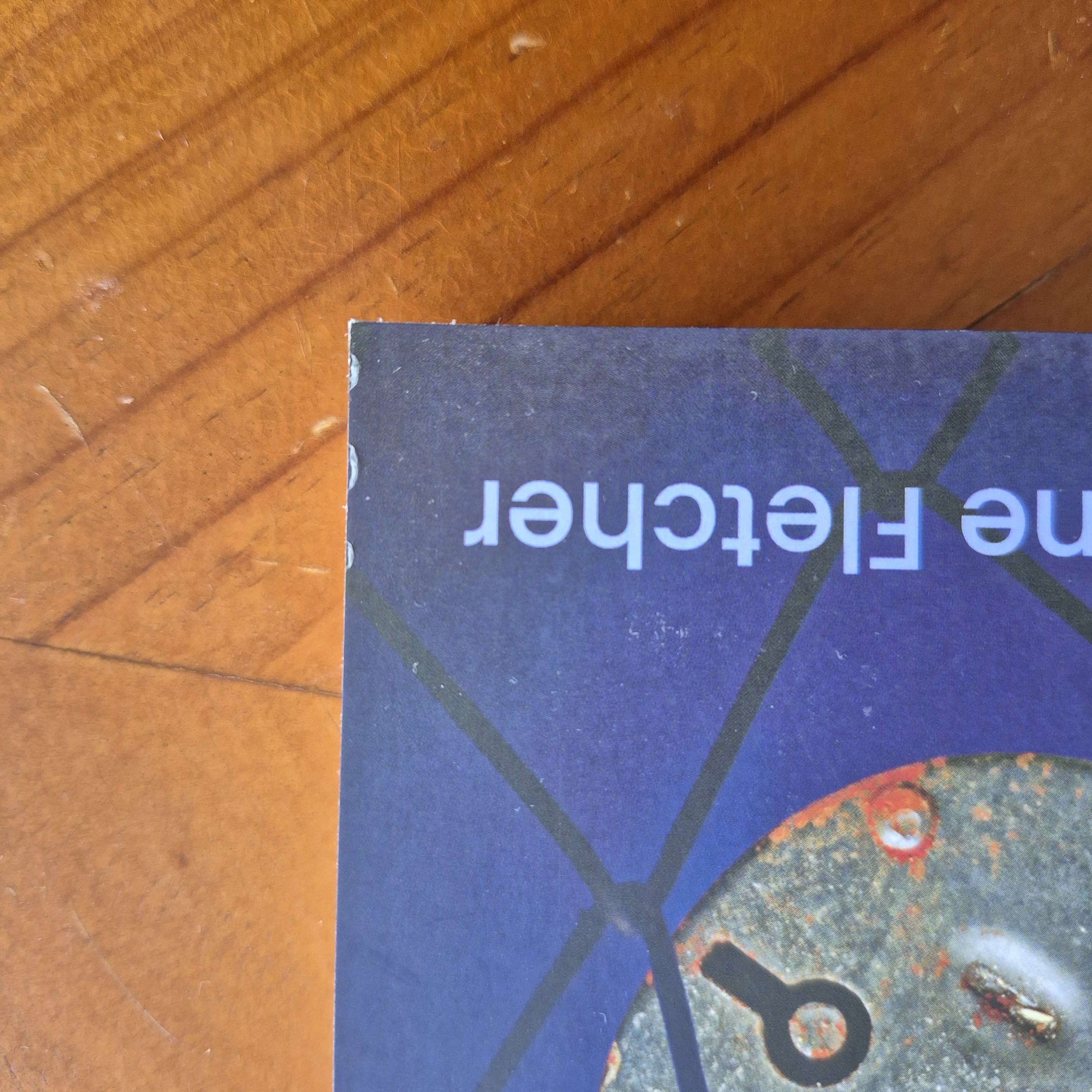 The corner of a blue book on the wooden table. The book corner has slight damage where the laminate is pulling away from the card.