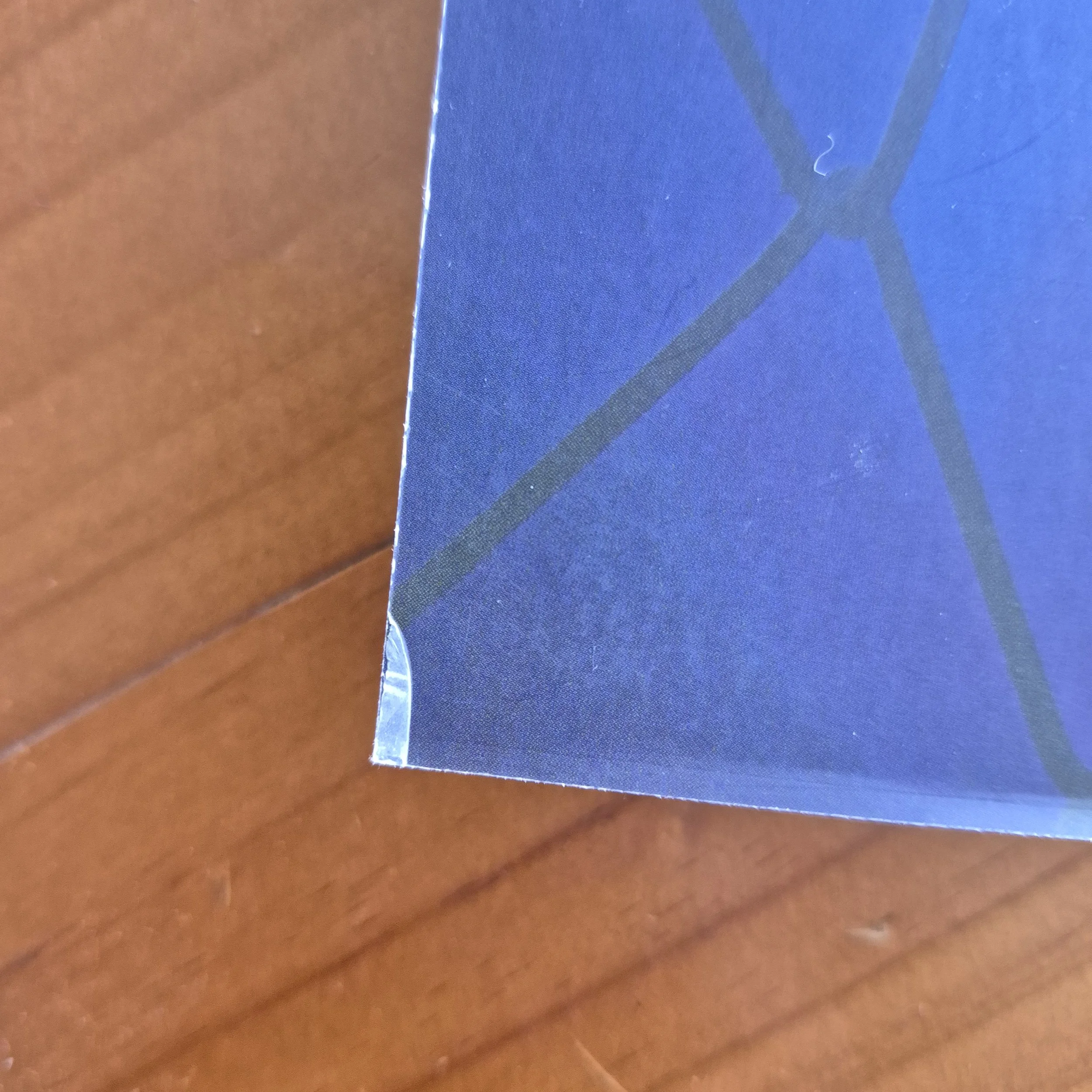 The corner of a blue book on the wooden table. The book corner has slight damage where the laminate is pulling away from the card.
