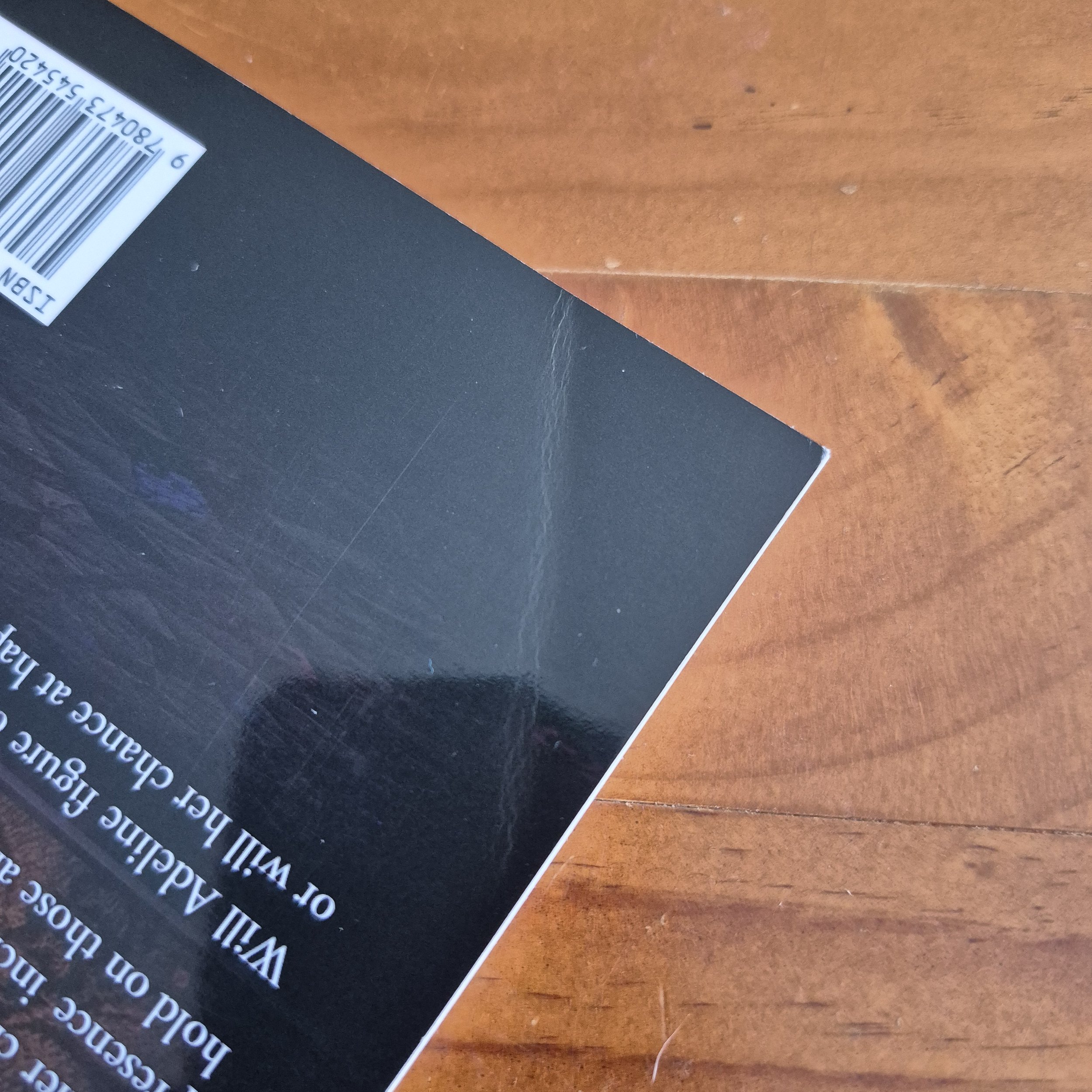 The corner of a book against a wooden background. The back cover is bent slightly, leaving a line near the corner.