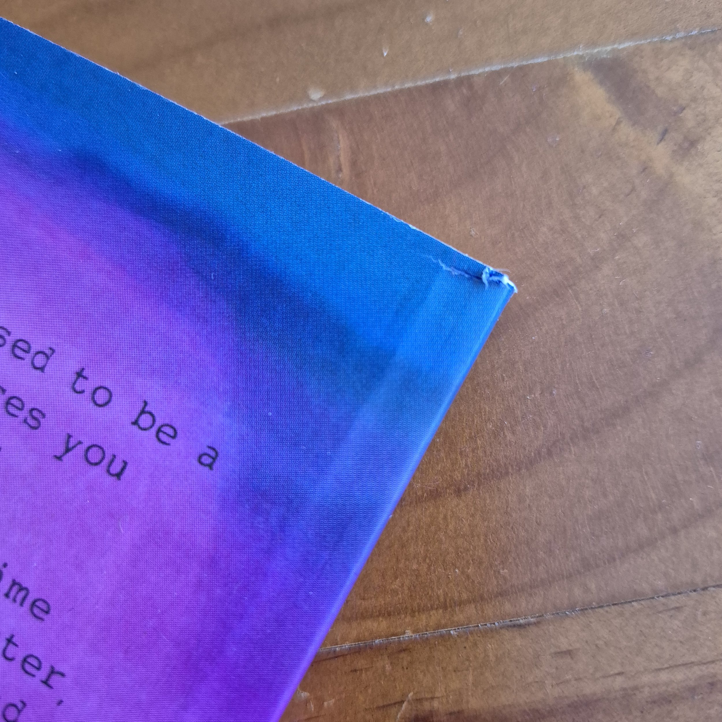 The corner of a book against a wooden background. The cover has a tiny tear near the spine on the back.