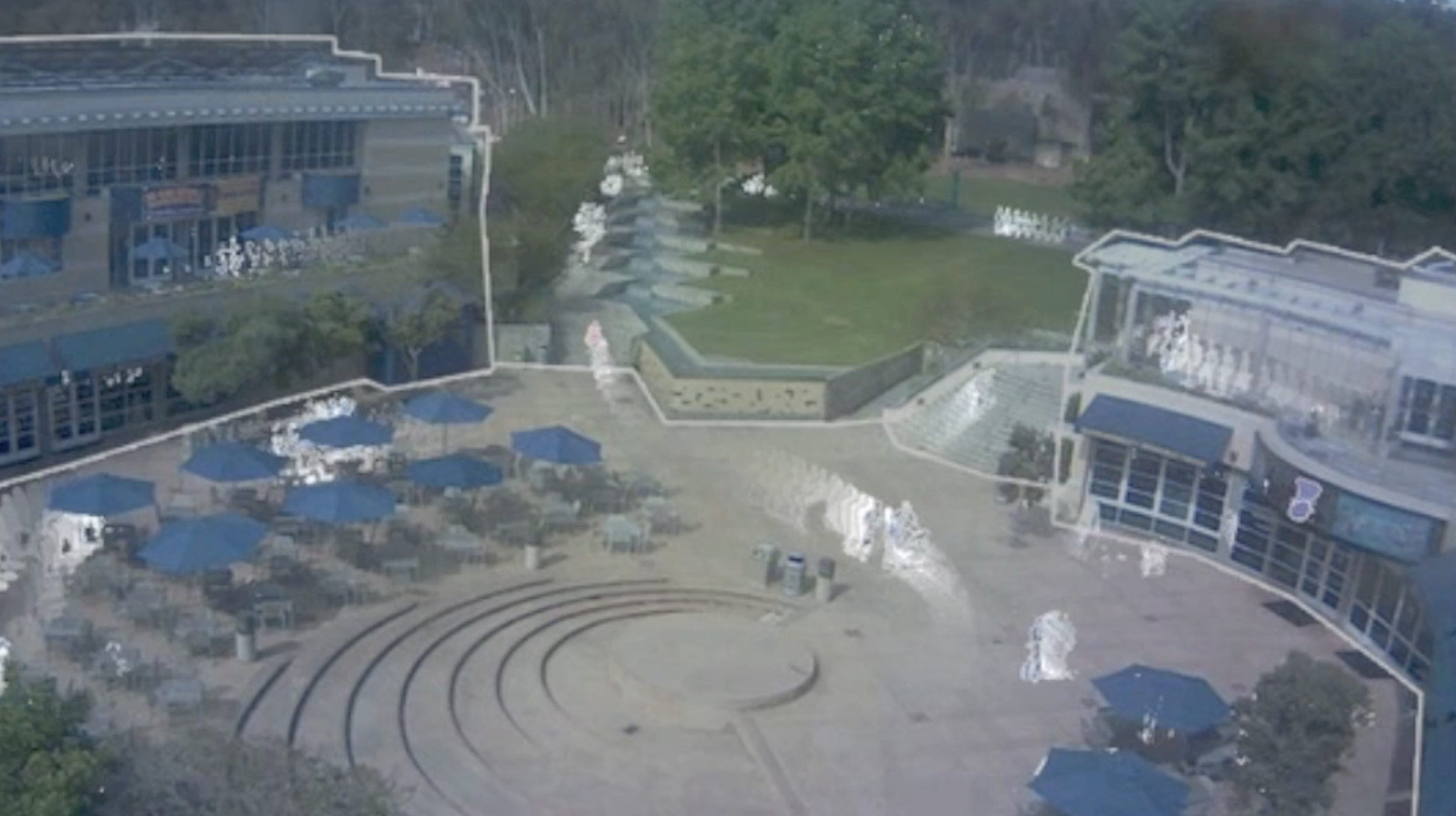 Aerial view of a shopping mall or commercial area with outdoor seating, umbrellas, and a central stage. Two large buildings with glass facades and a green lawn are visible, surrounded by trees.