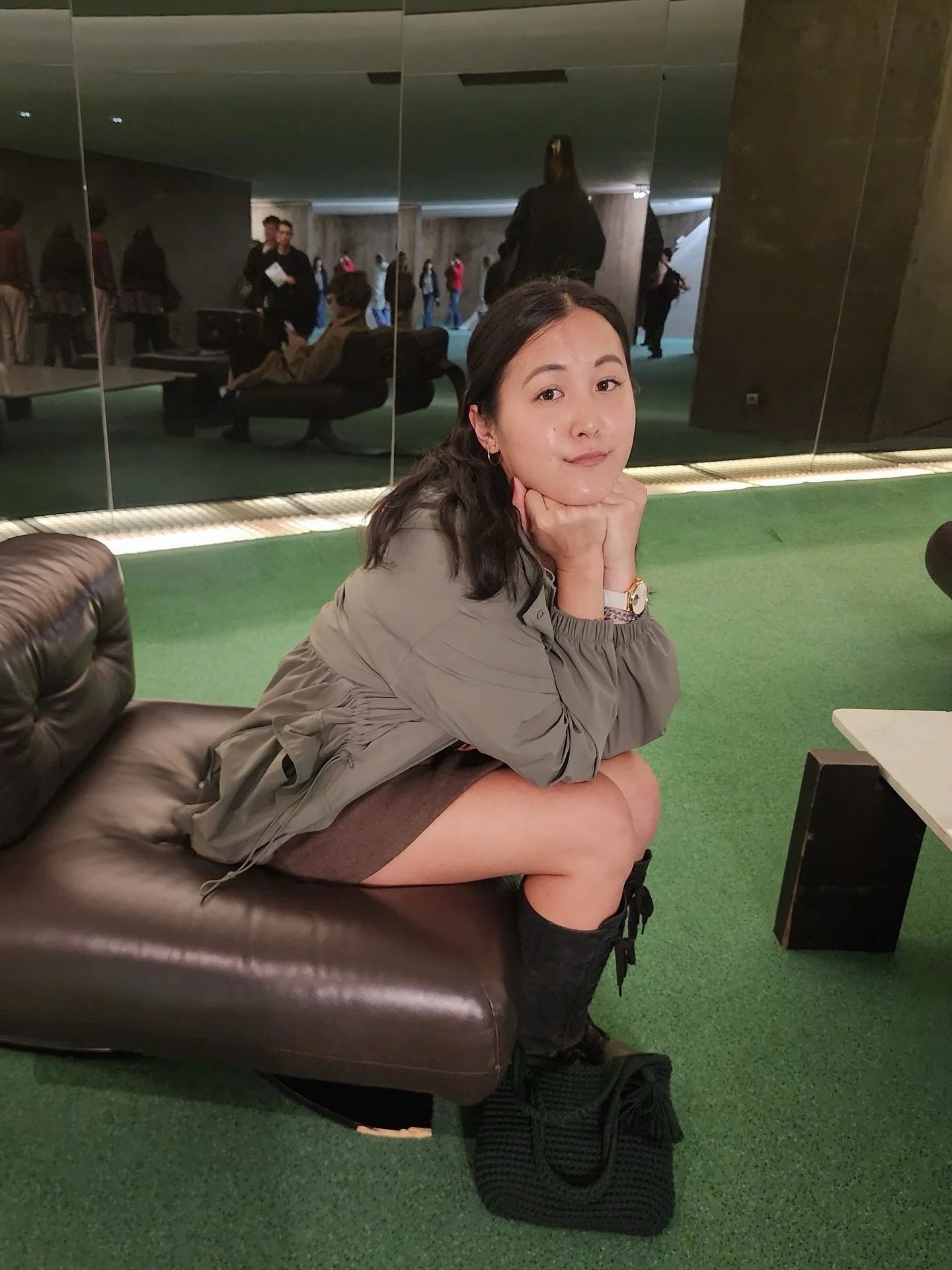 A young woman with dark hair sitting on a leather cushion, resting her chin on her hands, in an indoor setting with a green carpet. There are people in the background through a large mirror, suggesting a public or waiting area.