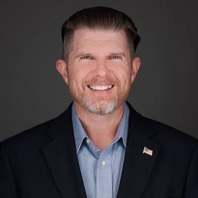 Smiling man in a suit with an American flag lapel pin on a dark background.