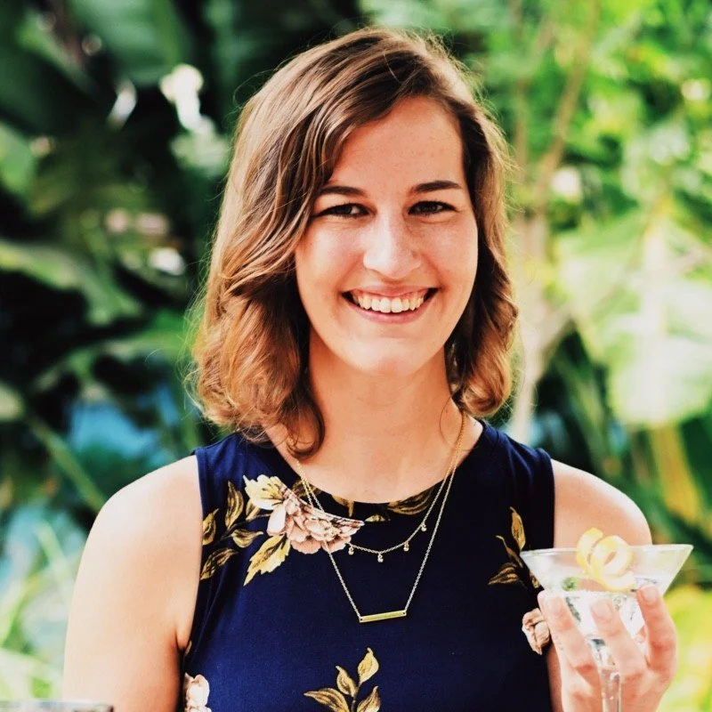 Smiling woman holding a cocktail in a garden setting, wearing a floral dress