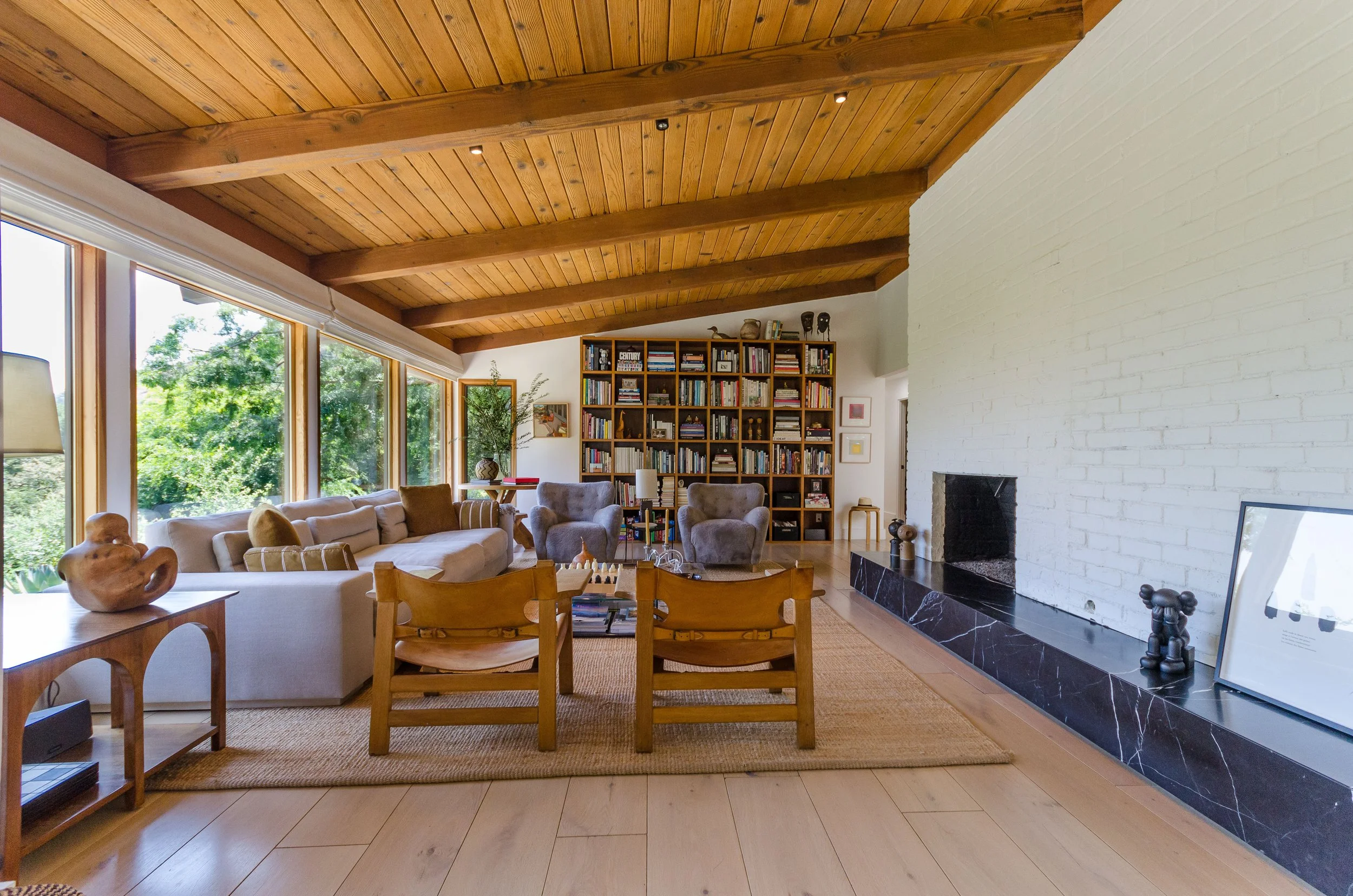 Living room with wooden ceiling, large windows, white brick fireplace, and a bookshelf filled with books.