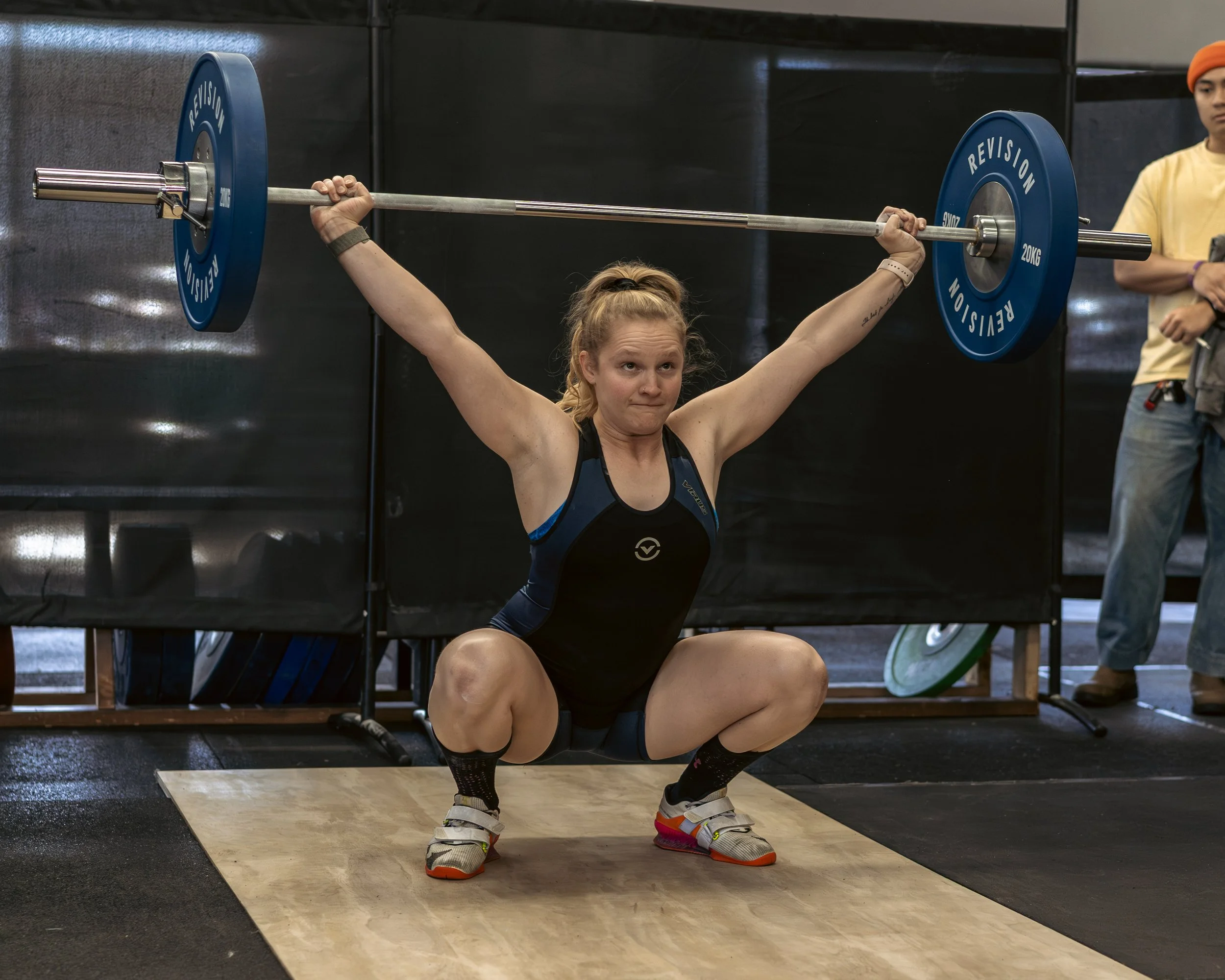 A woman with blonde hair in a bun is performing a workout, squatting with a barbell on her shoulders in a gym. She is wearing a black athletic outfit and colorful sneakers. Another person in a yellow shirt and orange beanie is standing nearby, observing.