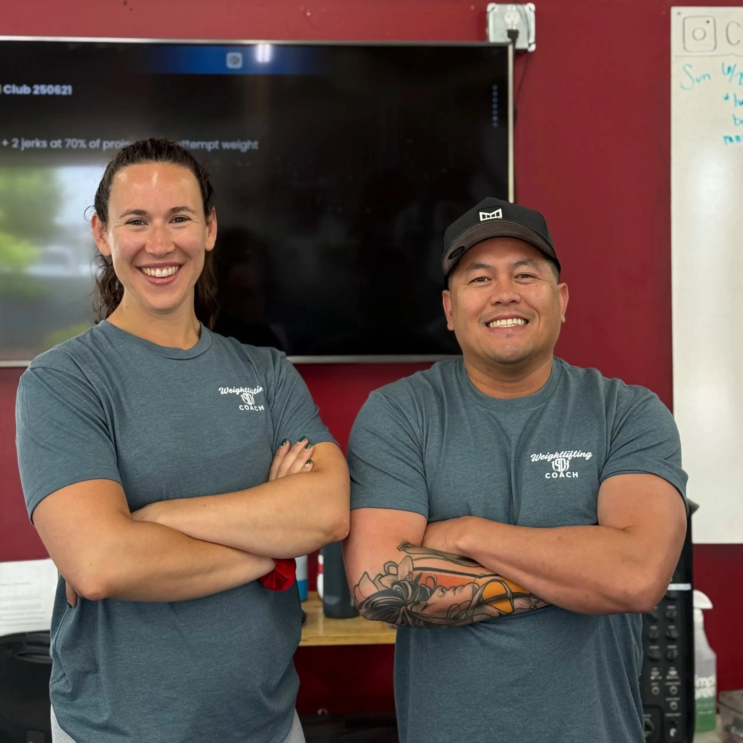 Two people, a woman and a man, smiling with arms crossed, standing in front of a black screen in an indoor setting. The woman wears a gray T-shirt with white lettering, and the man wears a gray T-shirt with white lettering and a black cap with a logo. There are business-related items in the background, including a whiteboard and shelves with snack packages.