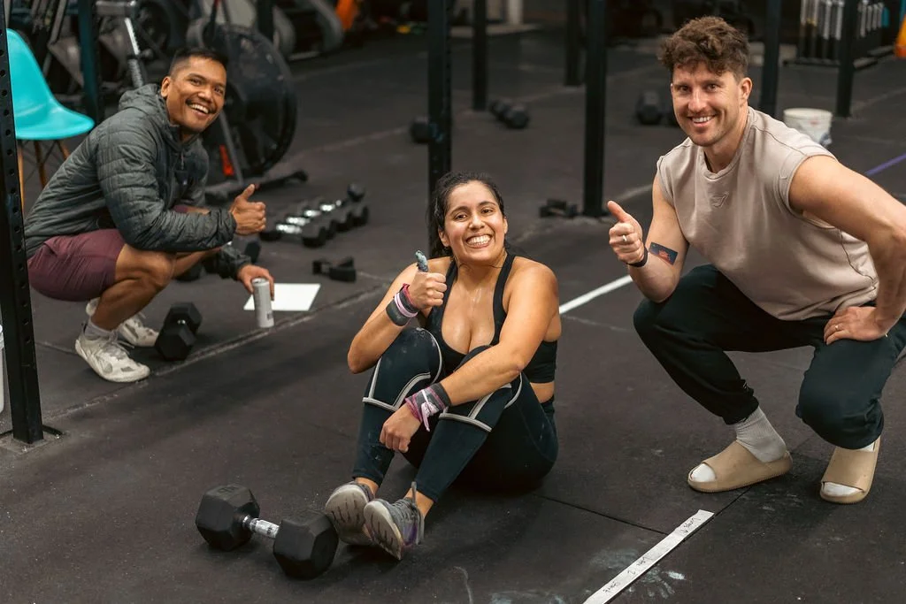 Three smiling people at a gym: a woman sitting on the floor with dumbbells, holding a thumbs-up, and two men squatting nearby, all giving thumbs-up.