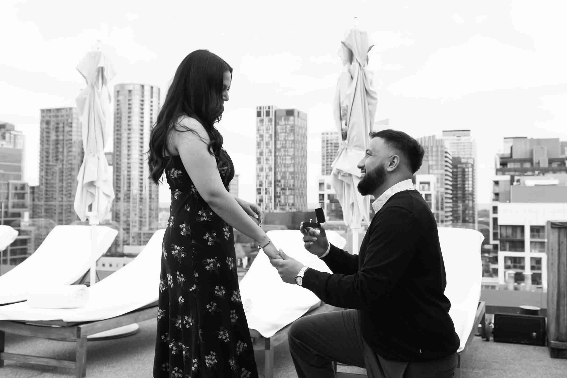 Groom on one knee proposing to a girl on a rooftop in downtown Toronto.