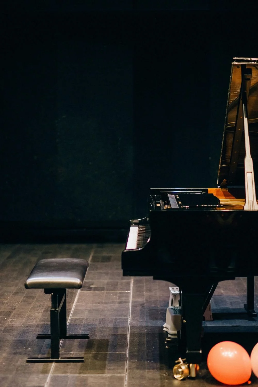 A black baby grand piano and bench on stage with black backdrop and orange balloon next to leg