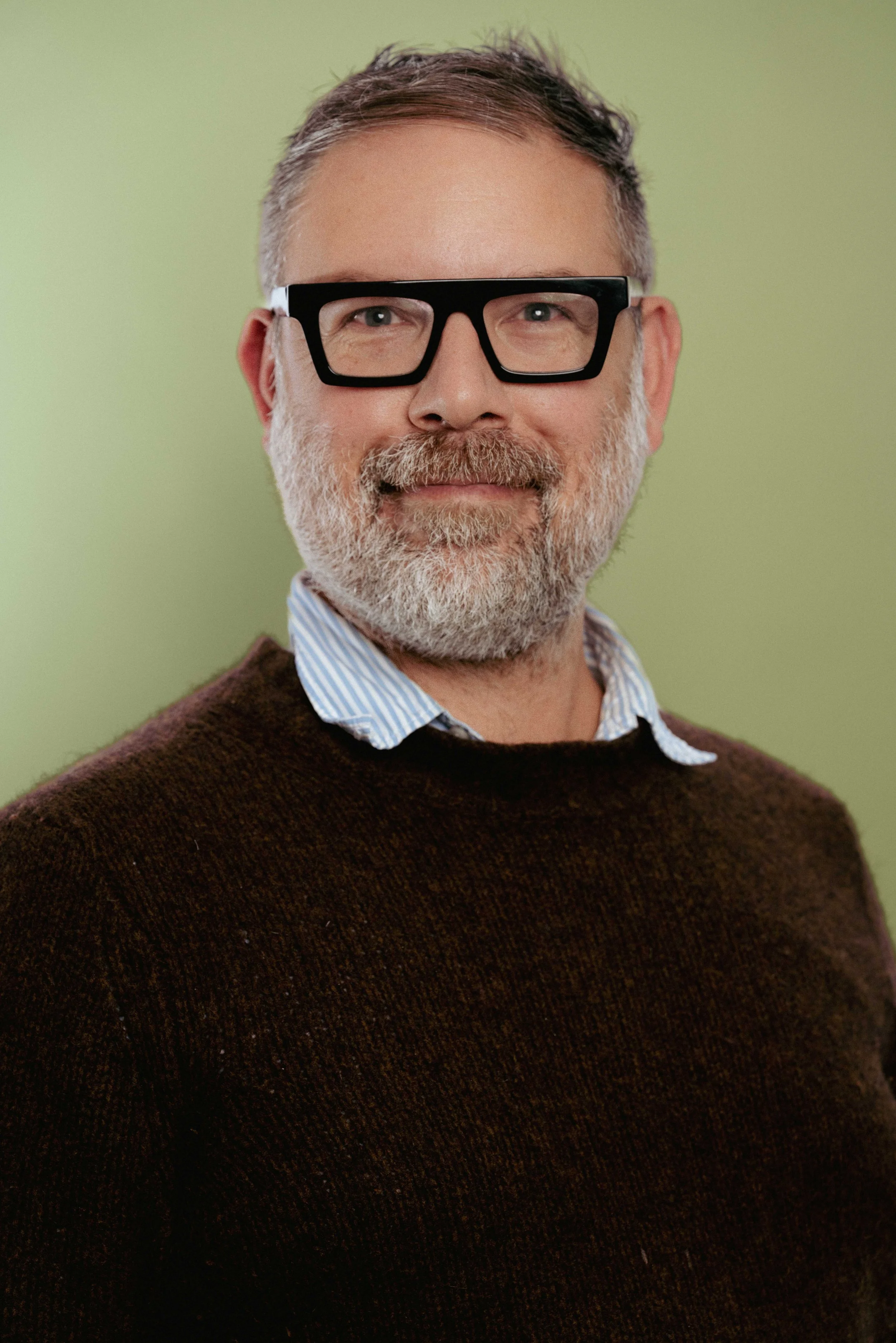 Man with glasses and beard wearing a dark sweater and button-up shirt at an event with logos in the background.