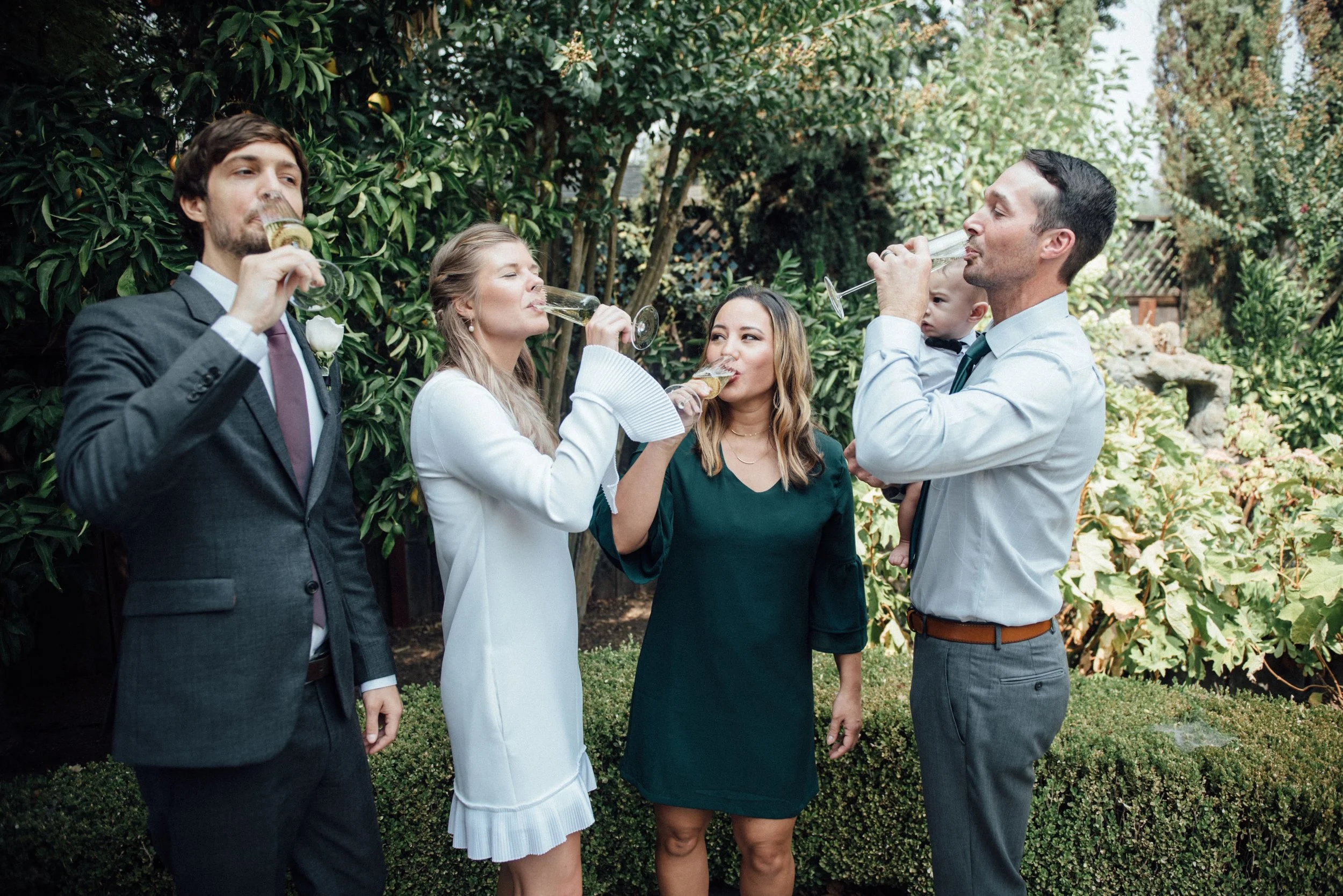 Five adults, dressed in formal attire, standing outdoors among greenery, drinking champagne from glasses, with a baby held by a man on right side.