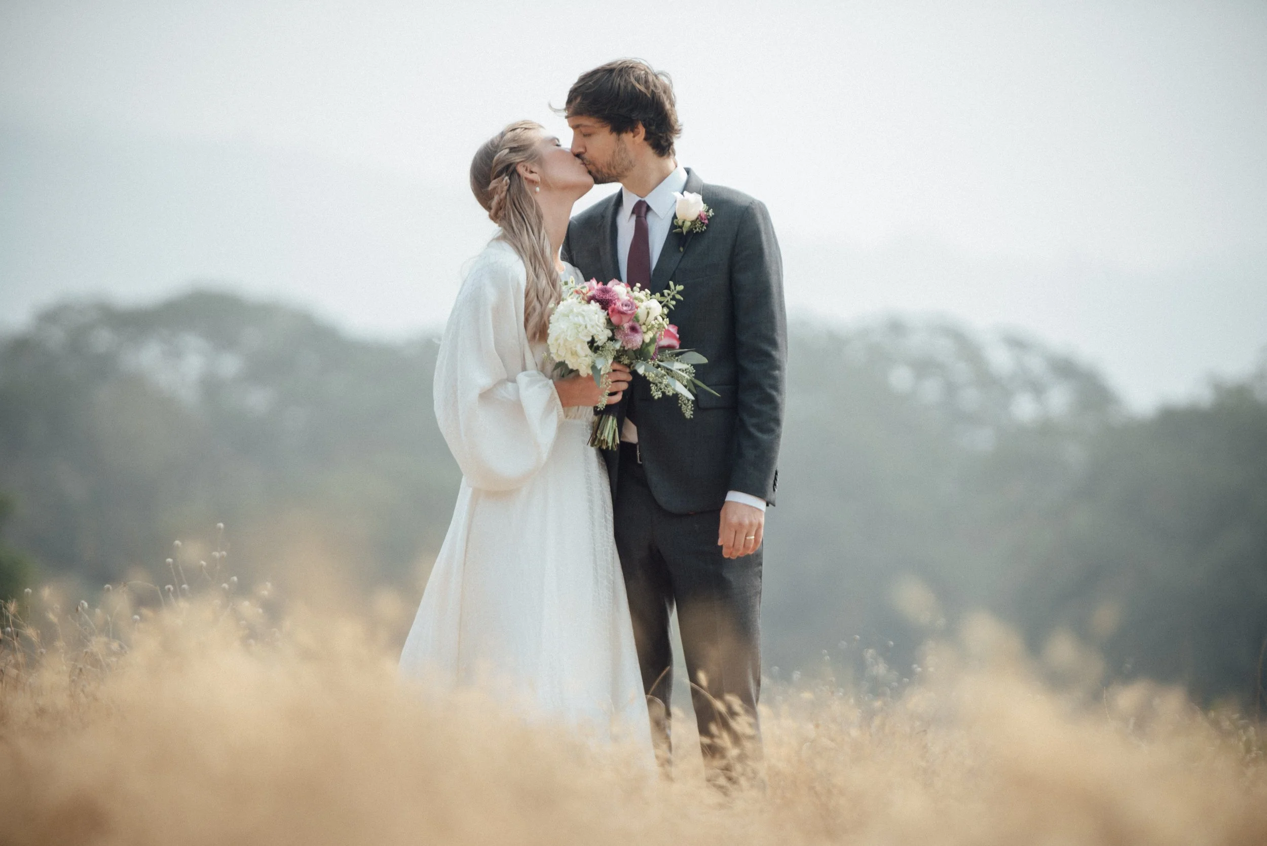 A bride and groom sharing a kiss outdoors, with the bride holding a bouquet of flowers, walking through a field of tall grass under a gray sky.