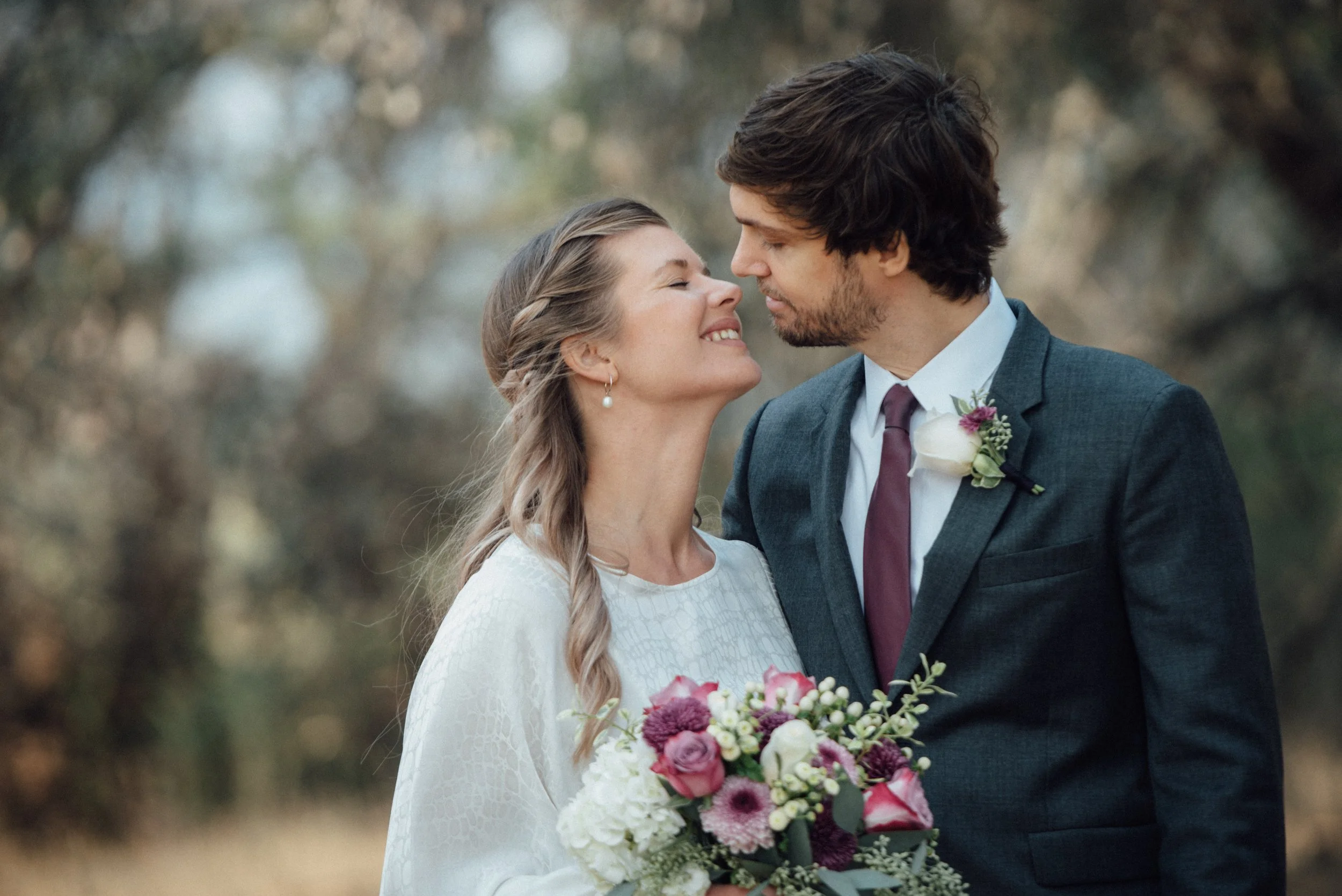 A bride and groom on their wedding day standing close, about to touch noses with eyes closed, outdoors with blurred trees in the background. The bride wears a white dress and holds a bouquet of pink and white flowers, and the groom wears a dark gray