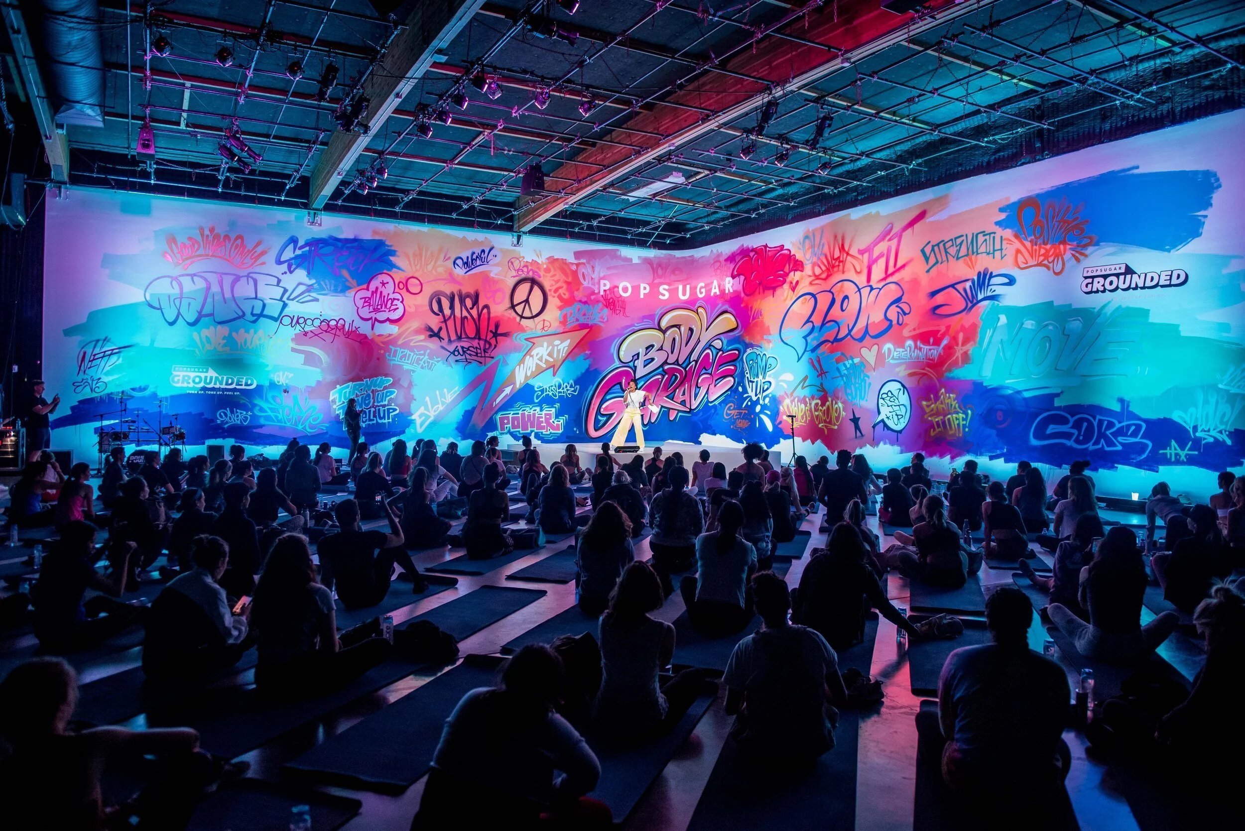pop sugar event. los angeles event photographer. A large indoor event with many people seated on yoga mats, listening to a speaker on stage in front of a colorful graffiti-style mural.