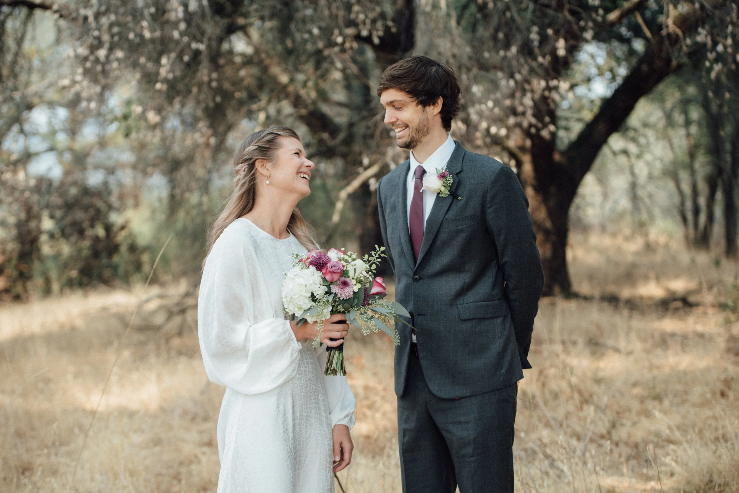 A bride and groom standing outdoors during a wedding, smiling at each other, with trees and dry grass in the background.