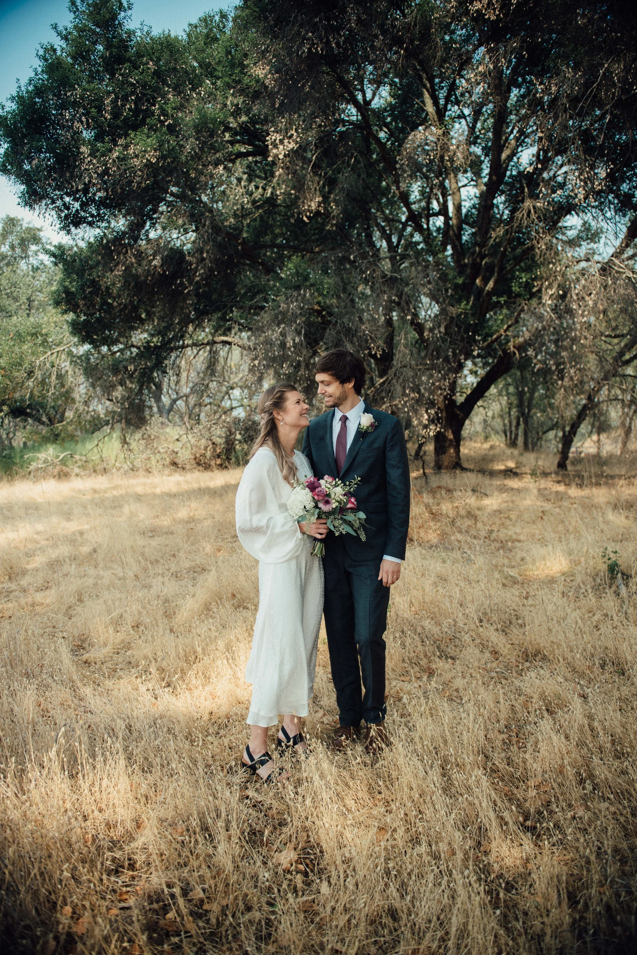 A bride and groom standing close together in a grassy field under a large tree during their wedding, both smiling and looking into each other's eyes. The bride holds a bouquet of flowers, and the groom is dressed in a dark suit with a tie.