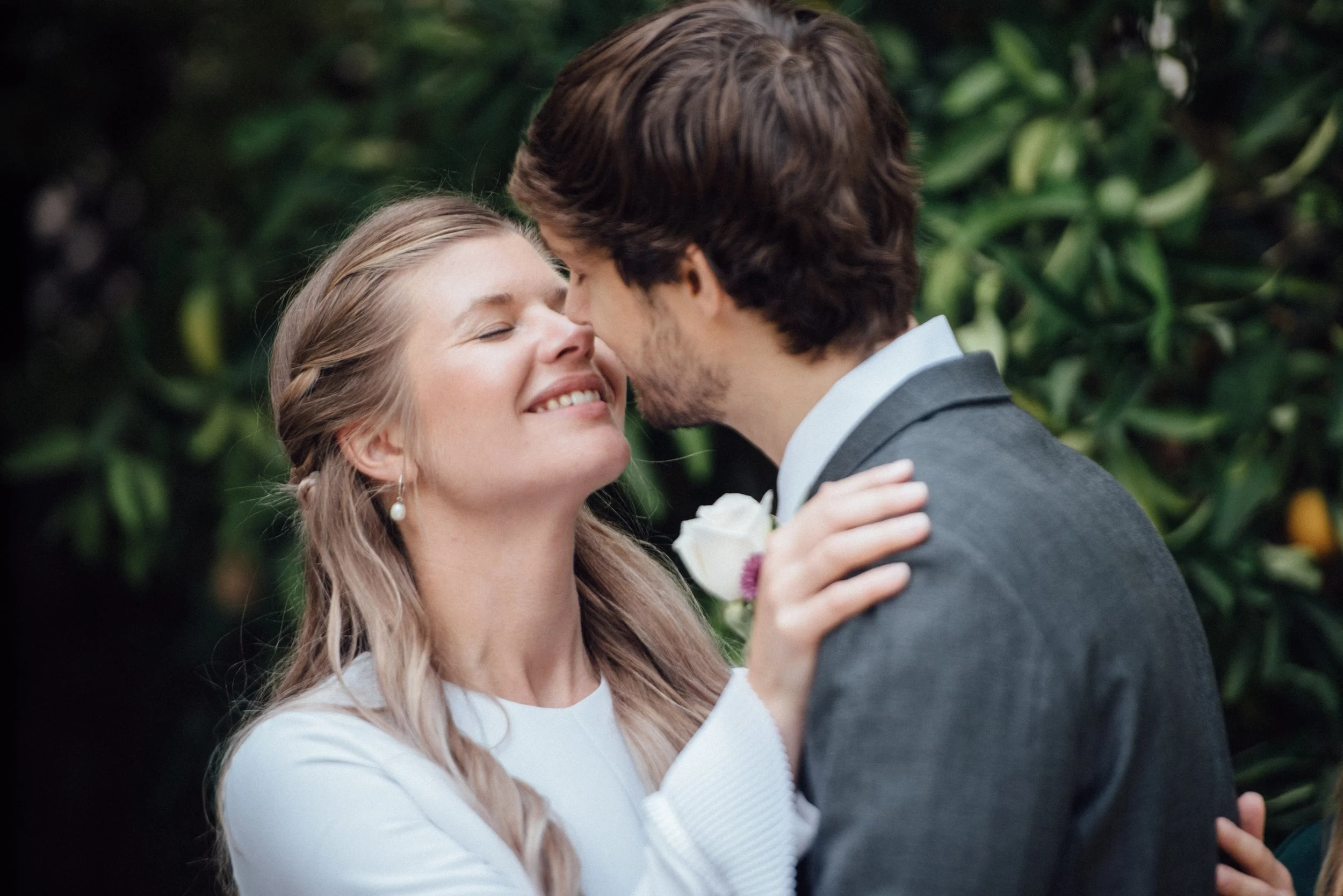 A smiling woman and a man embrace lovingly during a wedding ceremony outdoors, with green foliage in the background.