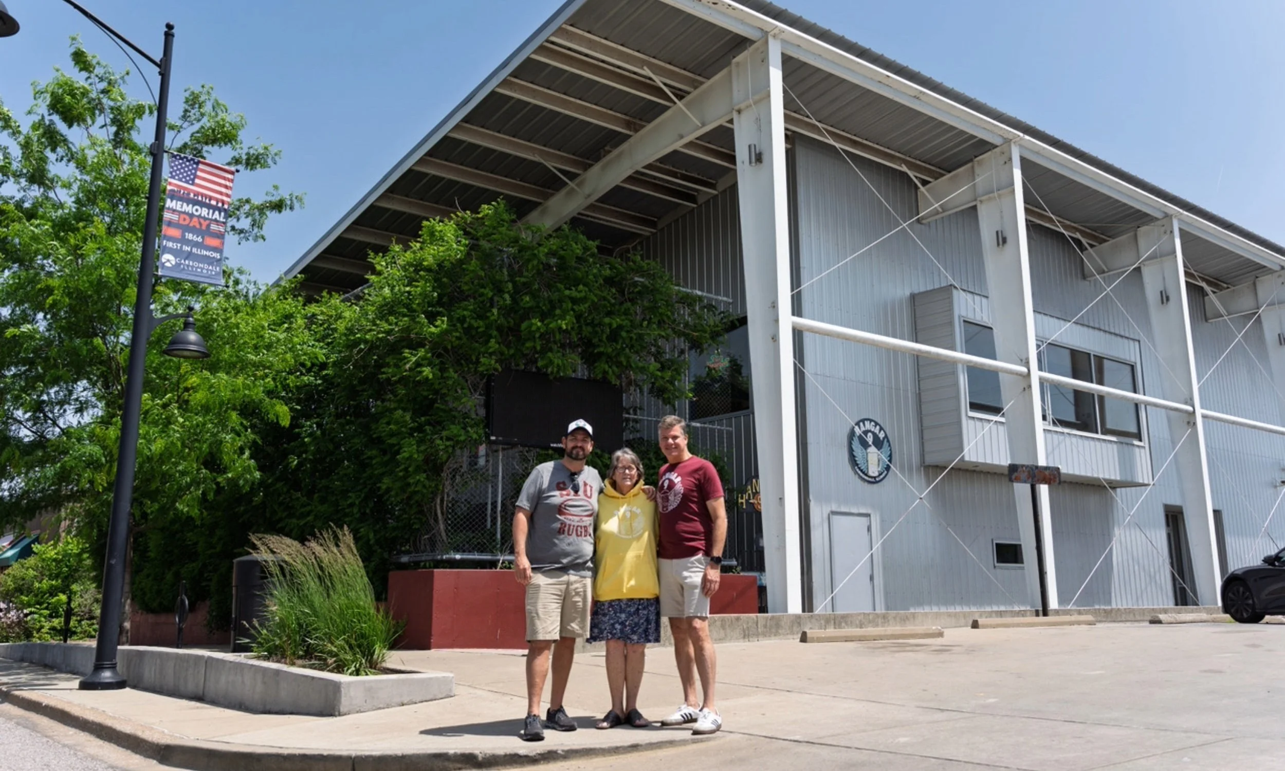 Three people standing in front of a building with a sign that reads 'Hangar' and a logo. One person is a woman wearing a yellow hoodie and a skirt, and two men are wearing casual clothes, standing on a sidewalk near a parking lot, with greenery.