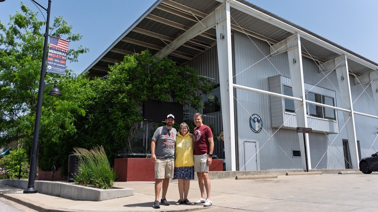 Three people standing in front of a building with a sign that reads 'Hangar' and a logo. One person is a woman wearing a yellow hoodie and a skirt, and two men are wearing casual clothes, standing on a sidewalk near a parking lot, with greenery and a lamppost nearby.