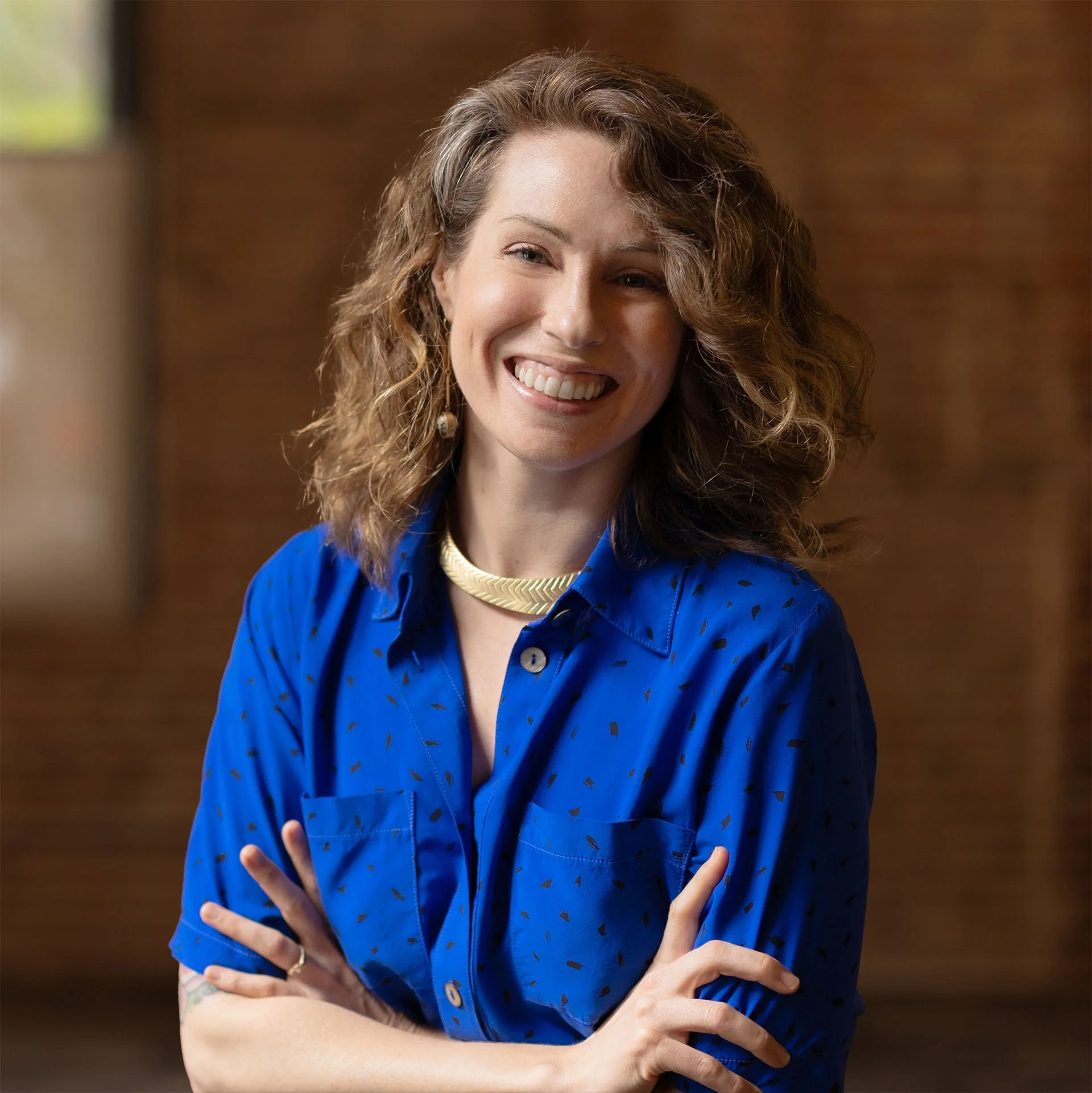 A woman with curly hair smiling and crossing her arms, wearing a blue blouse with a pattern, a gold necklace, and earrings, indoors with a brick wall background.