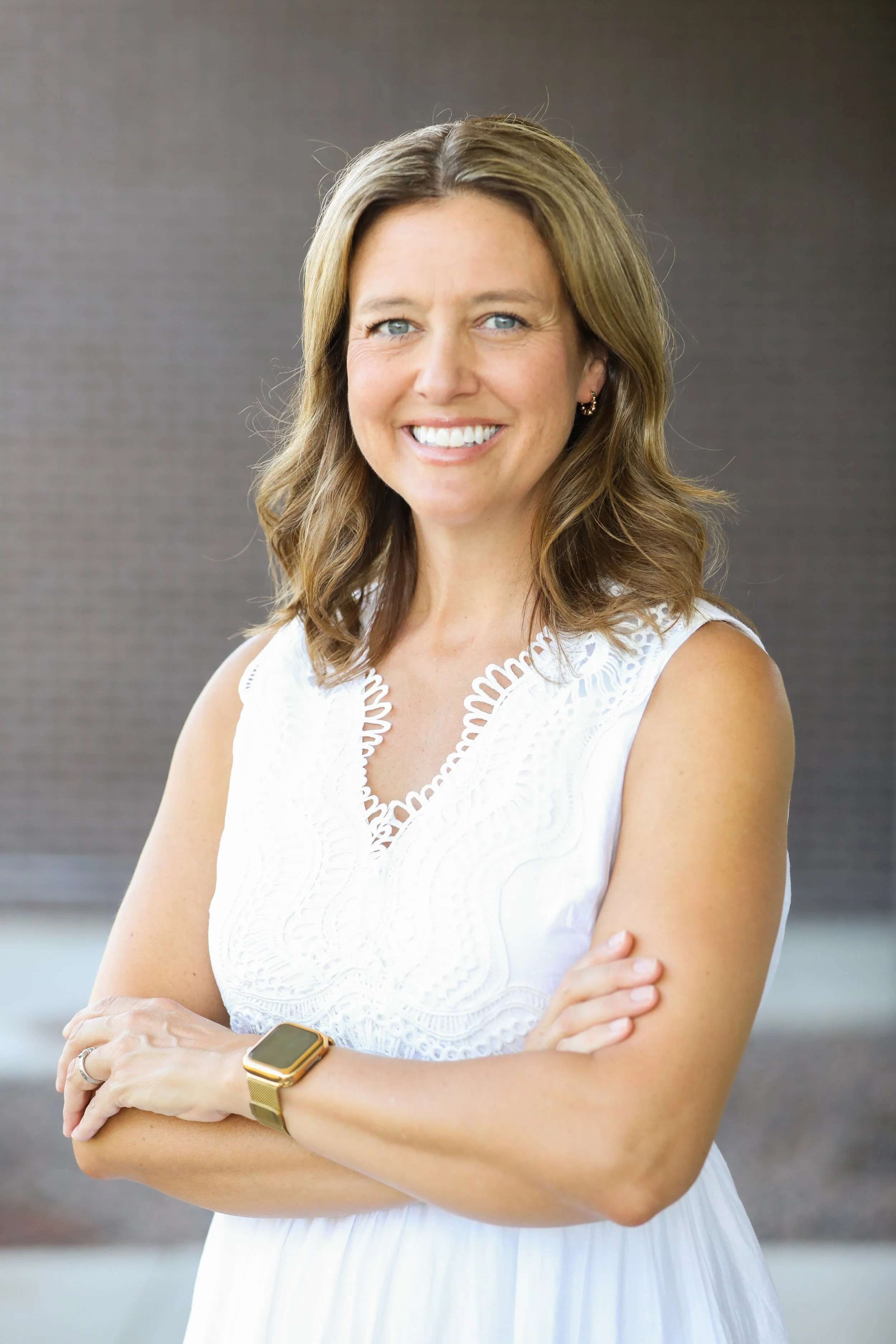 A smiling woman with shoulder-length light brown hair stands with her arms crossed, wearing a white sleeveless embroidered blouse