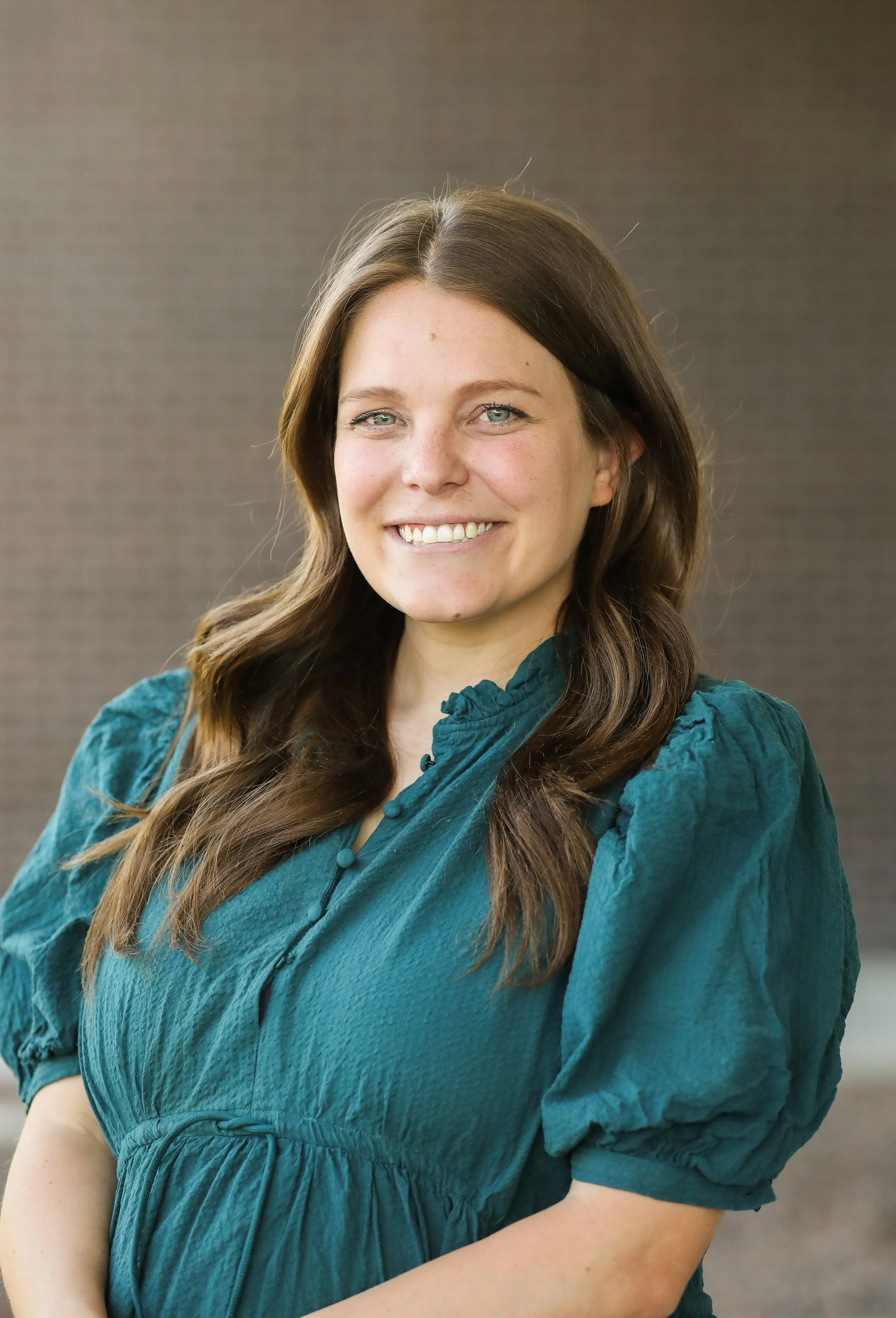 A smiling woman with long, wavy brown hair wears a teal dress