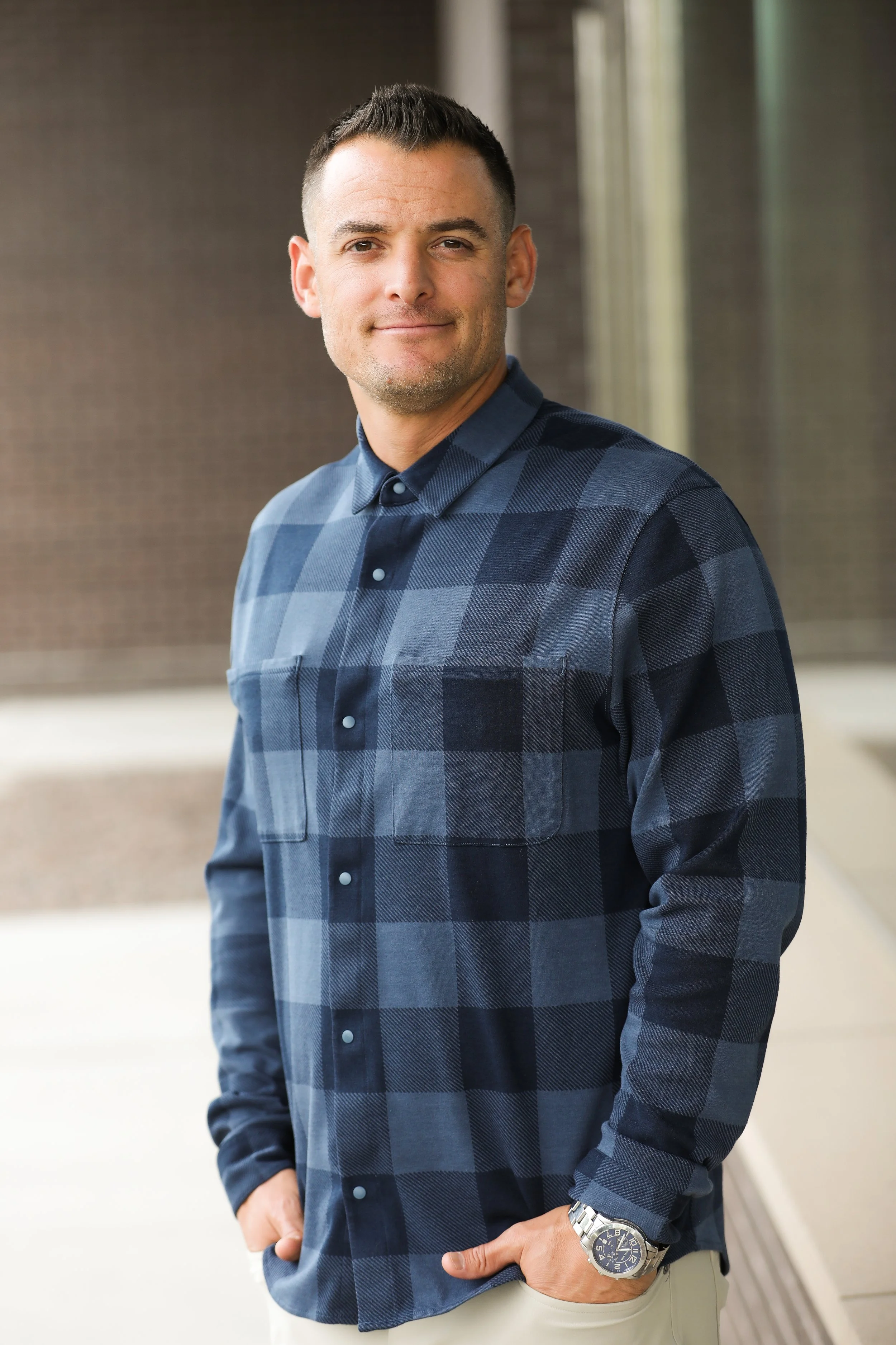 A man with short dark hair stands indoors, smiling at the camera while wearing a navy blue plaid button-up shirt