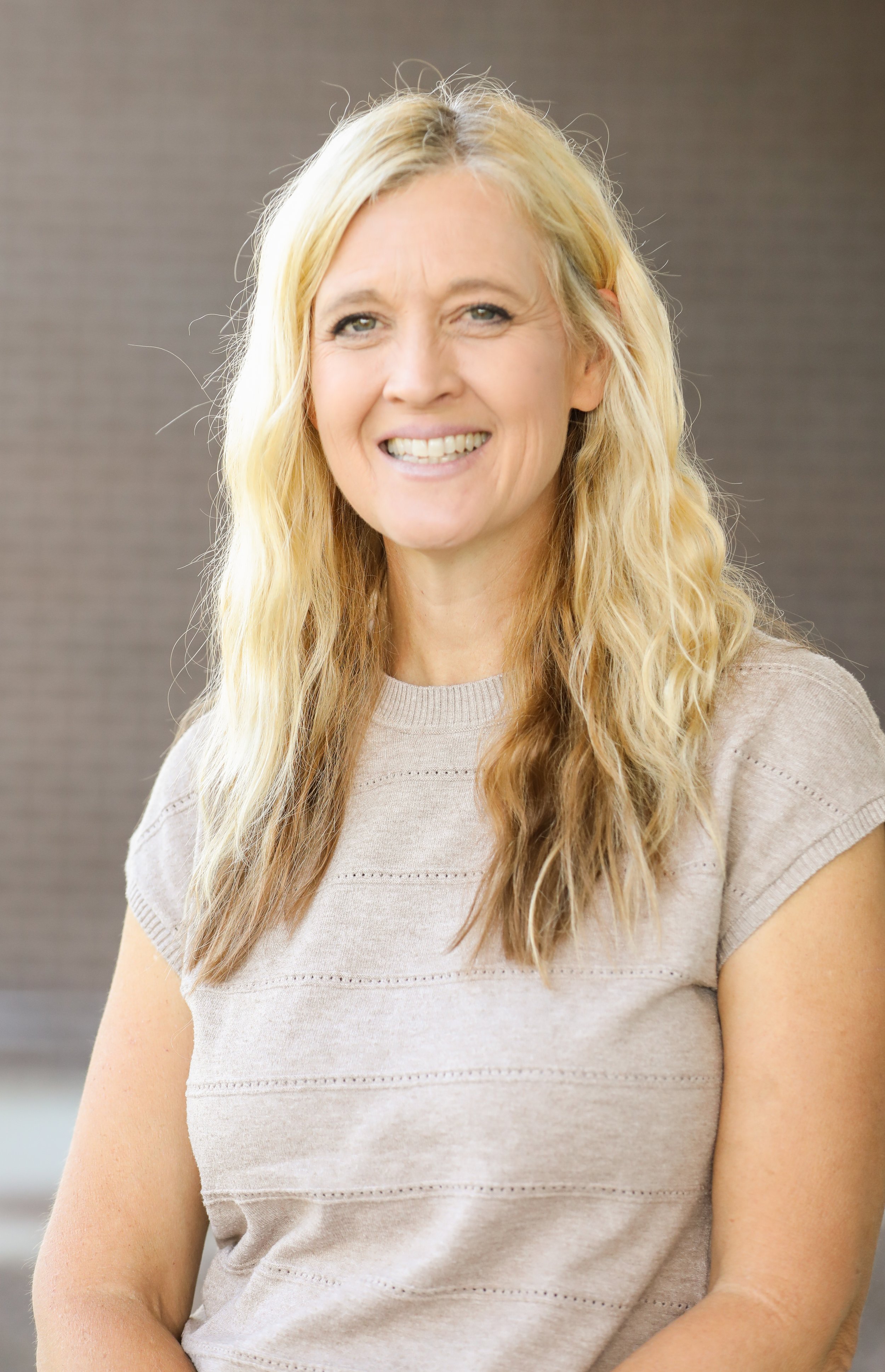 A smiling woman with long, wavy blonde hair wears a light beige knit top with short sleeves