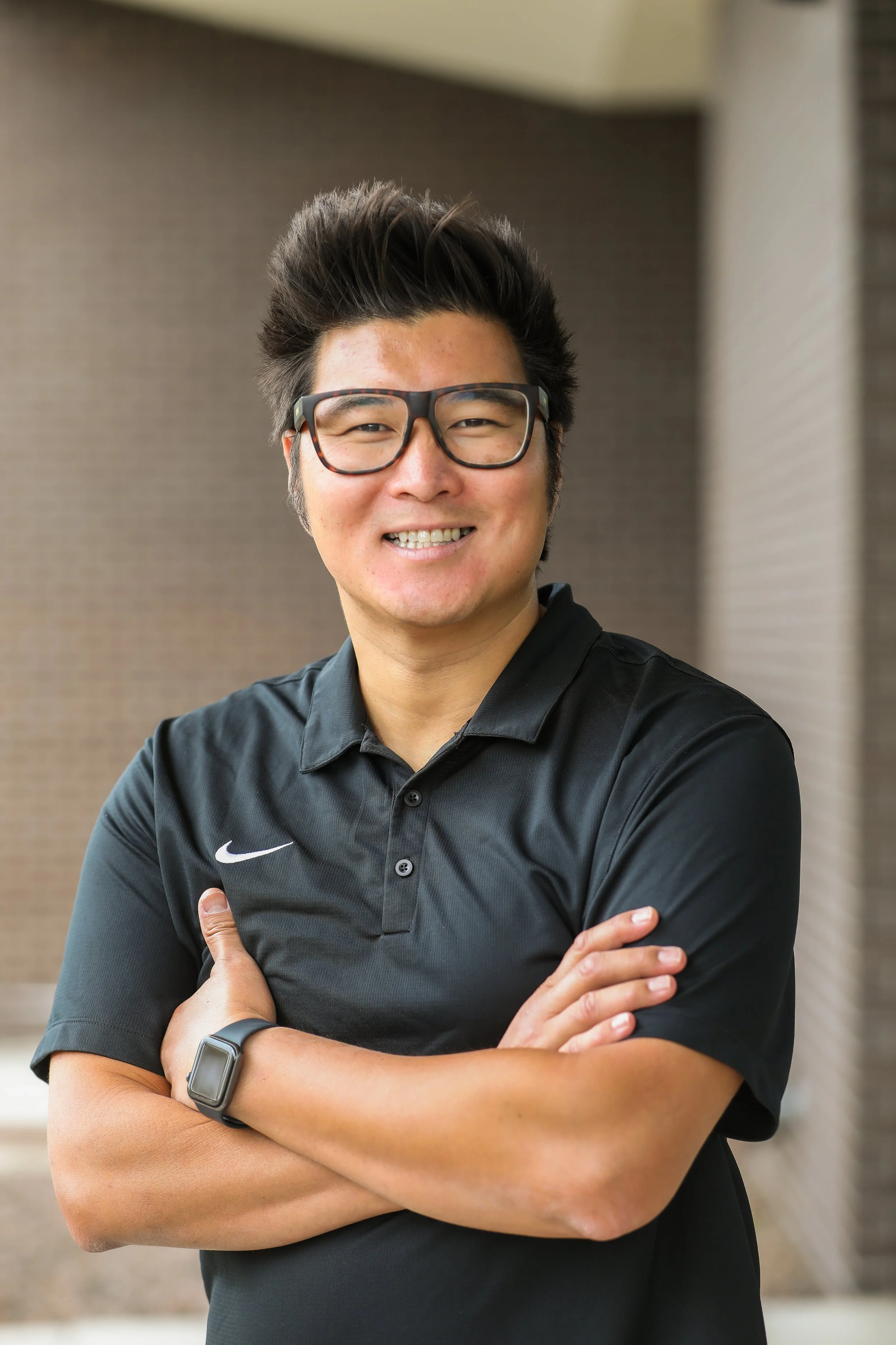 A smiling man wearing glasses and a black Nike polo shirt stands with his arms crossed, posing for a portrait indoors.