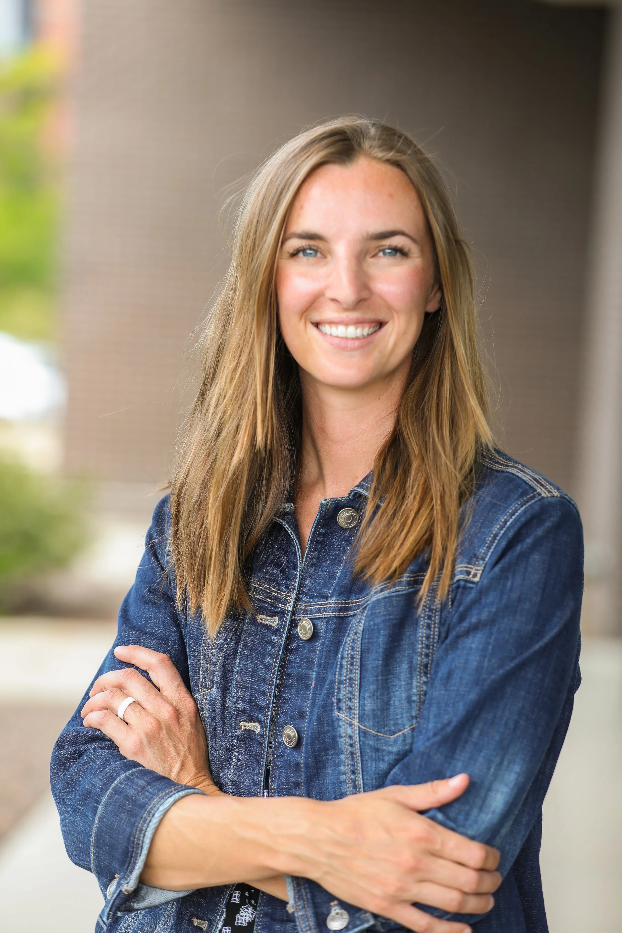 A smiling woman with long, light brown hair stands with her arms crossed, wearing a blue denim jacket