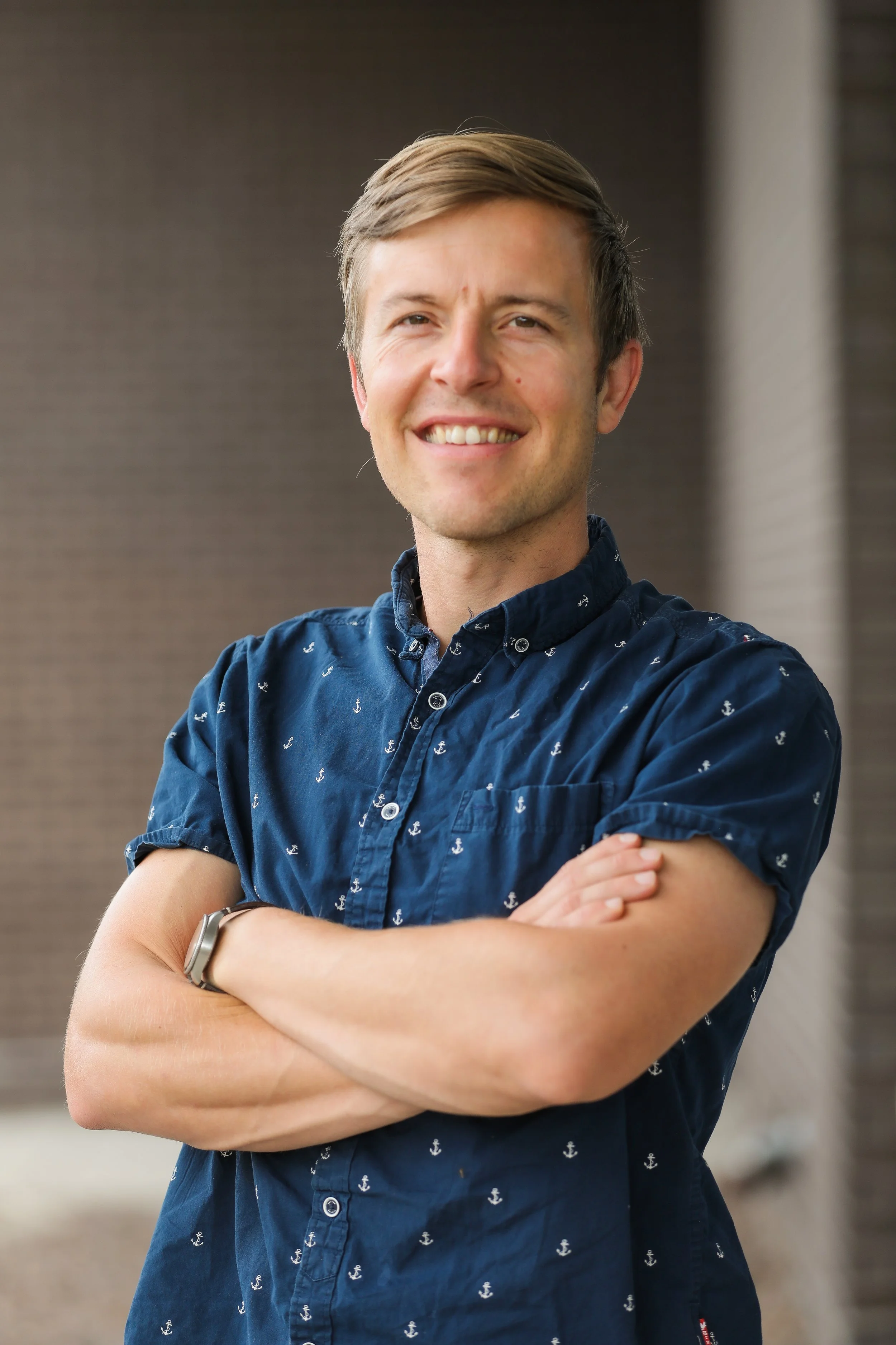 A smiling man with short, light brown hair stands with his arms crossed, wearing a navy blue button-up shirt