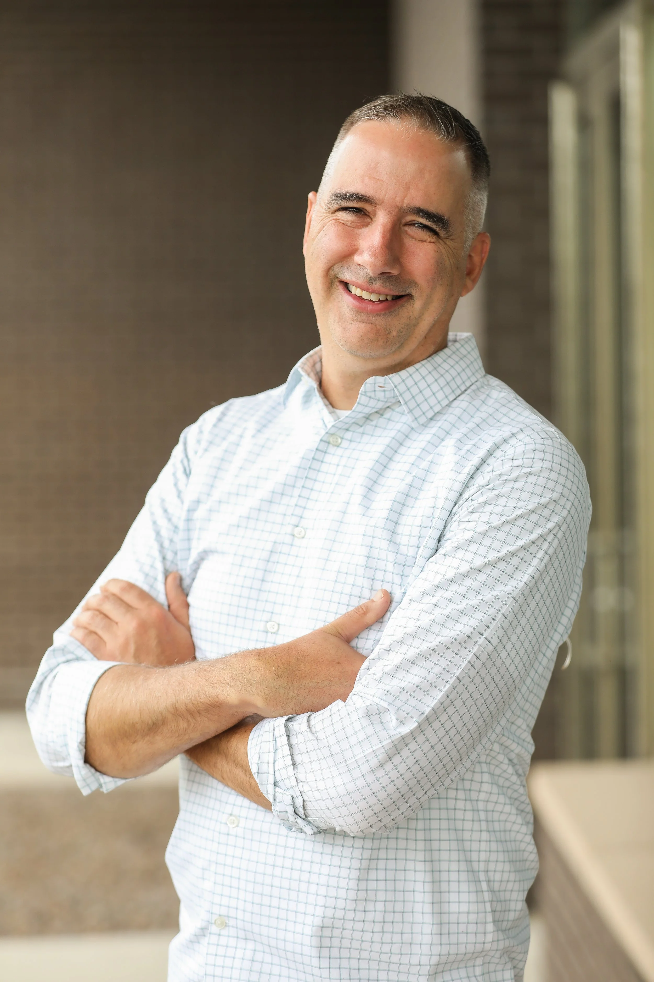 A smiling man with short gray hair stands with his arms crossed, wearing a white checkered button-up shirt. He poses outdoors in front of a modern building backdrop.