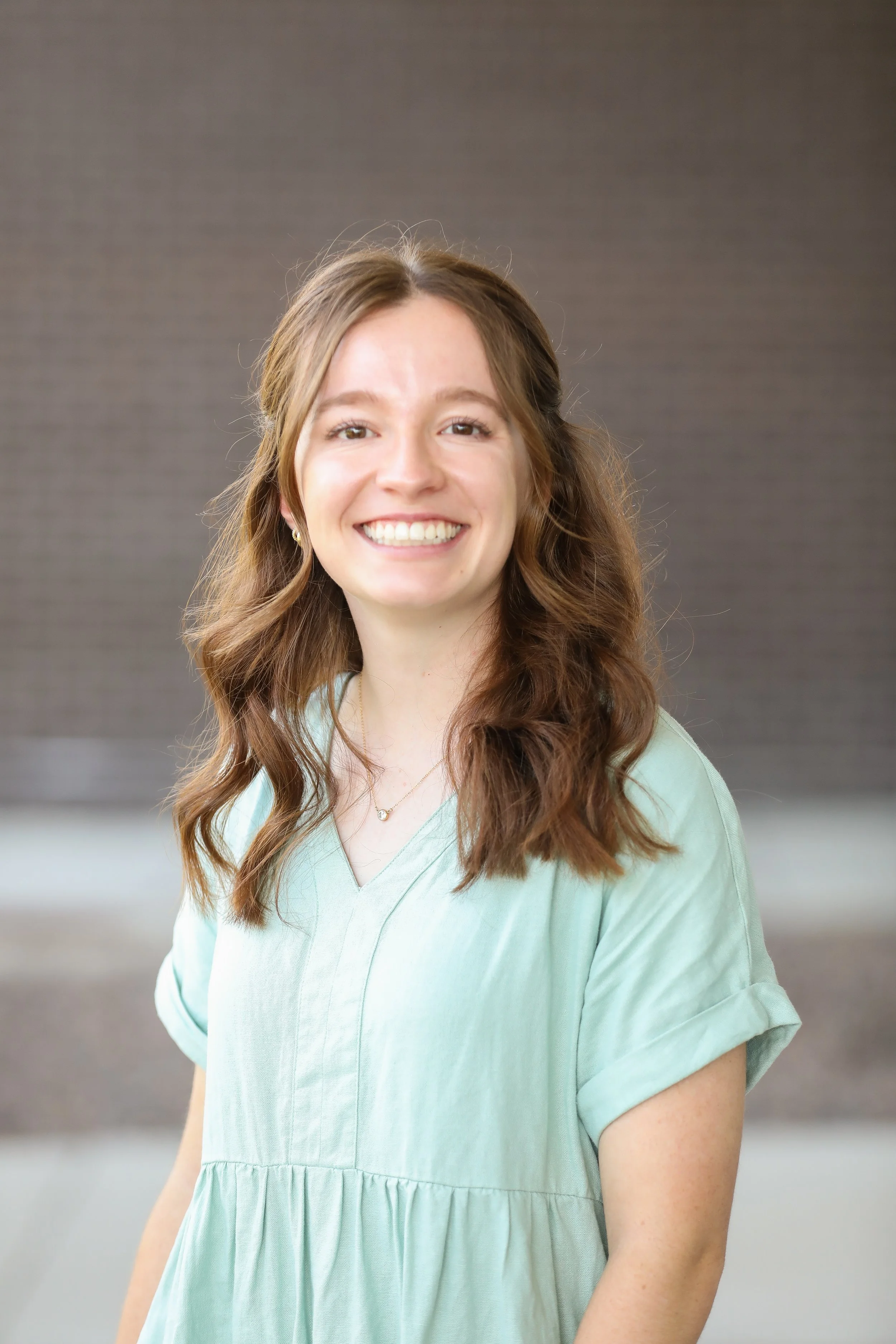 A smiling woman with long, wavy brown hair wears a light mint-green blouse with rolled sleeves and a delicate necklace