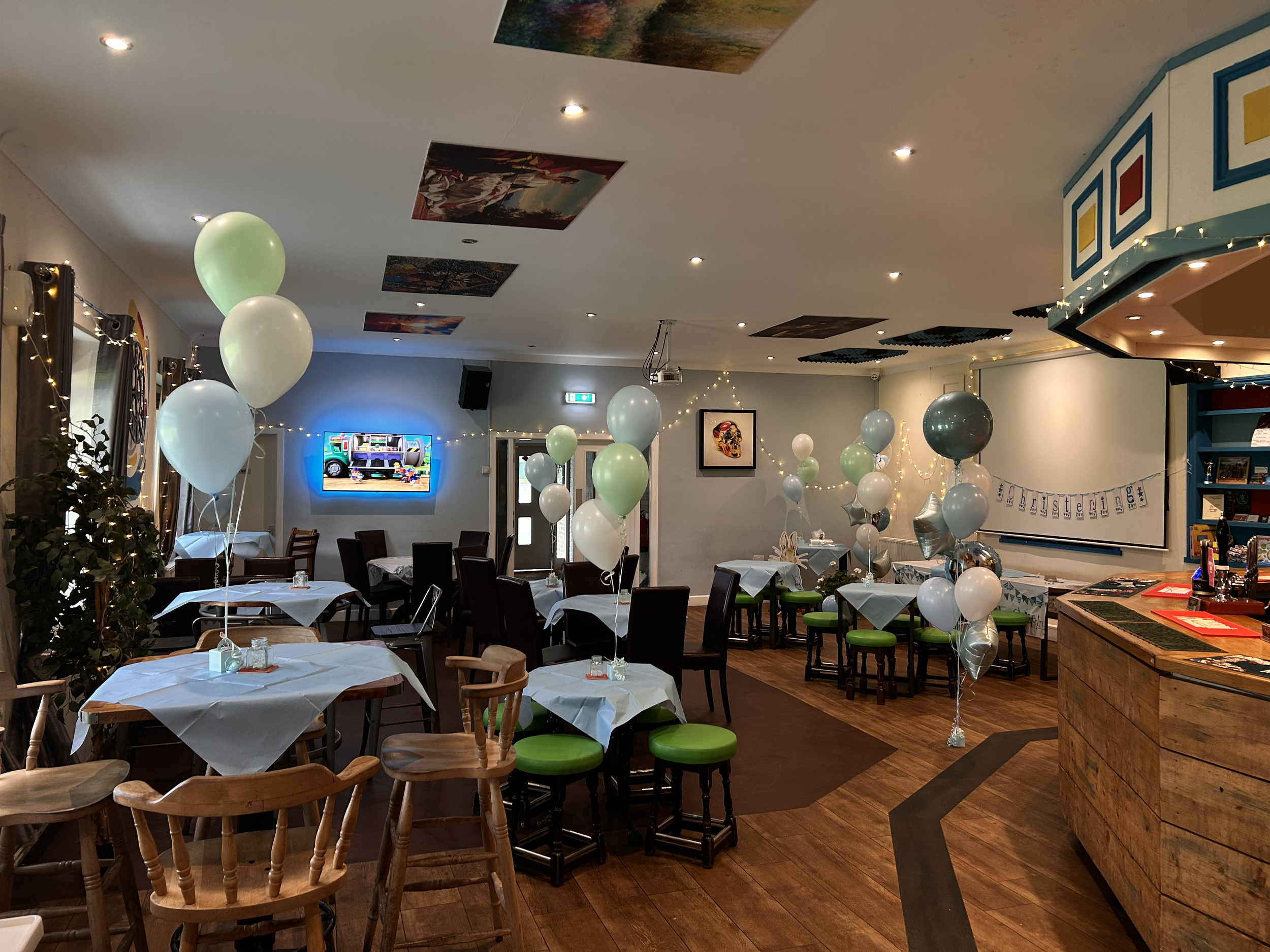 Decorated restaurant with tables covered in white tablecloths and green and white balloons, set for a celebration or party.