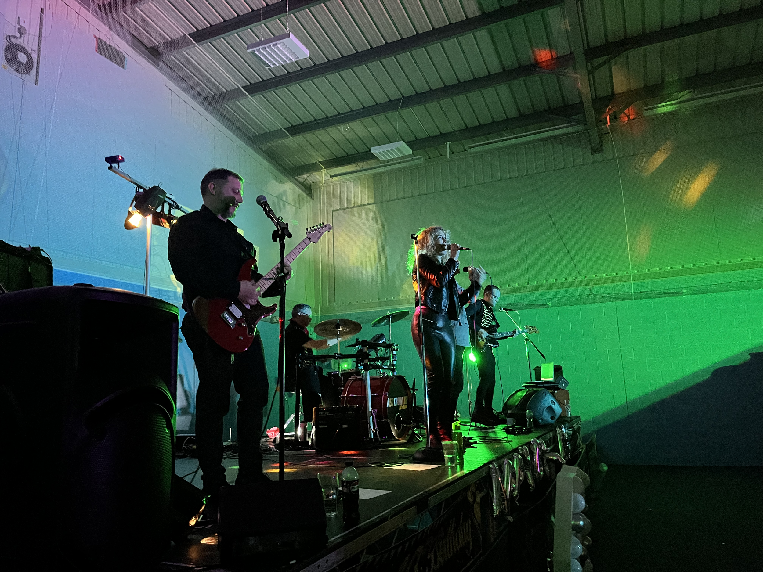 A band performing on stage with five members playing guitars, drums, and singing, in a large indoor venue with colorful lighting.