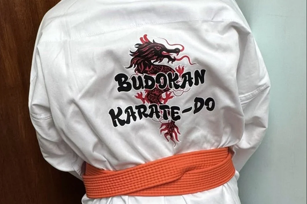Back of a white martial arts uniform with an embroidered dragon and the words "Budokan Karate-Do" on the back, paired with an orange belt.