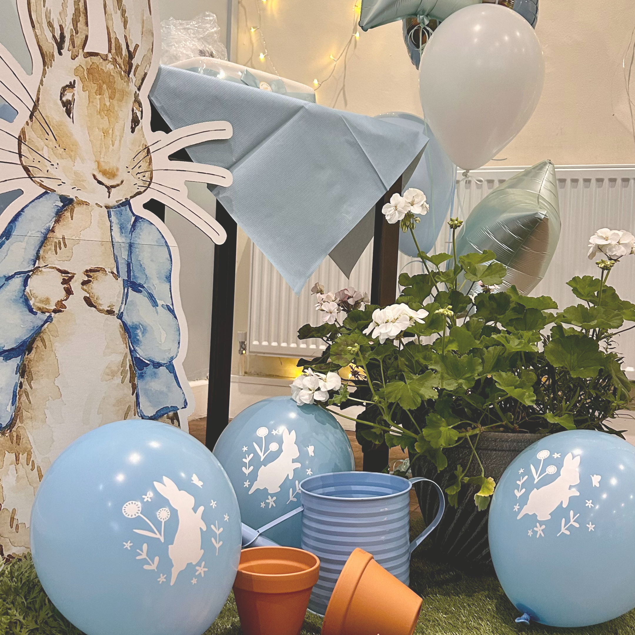 Easter display with blue balloons featuring bunny and floral designs, a potted plant with white flowers, a decorated table with a light blue tablecloth, a large cardboard bunny cutout, and scattered flowerpots, all set indoors.
