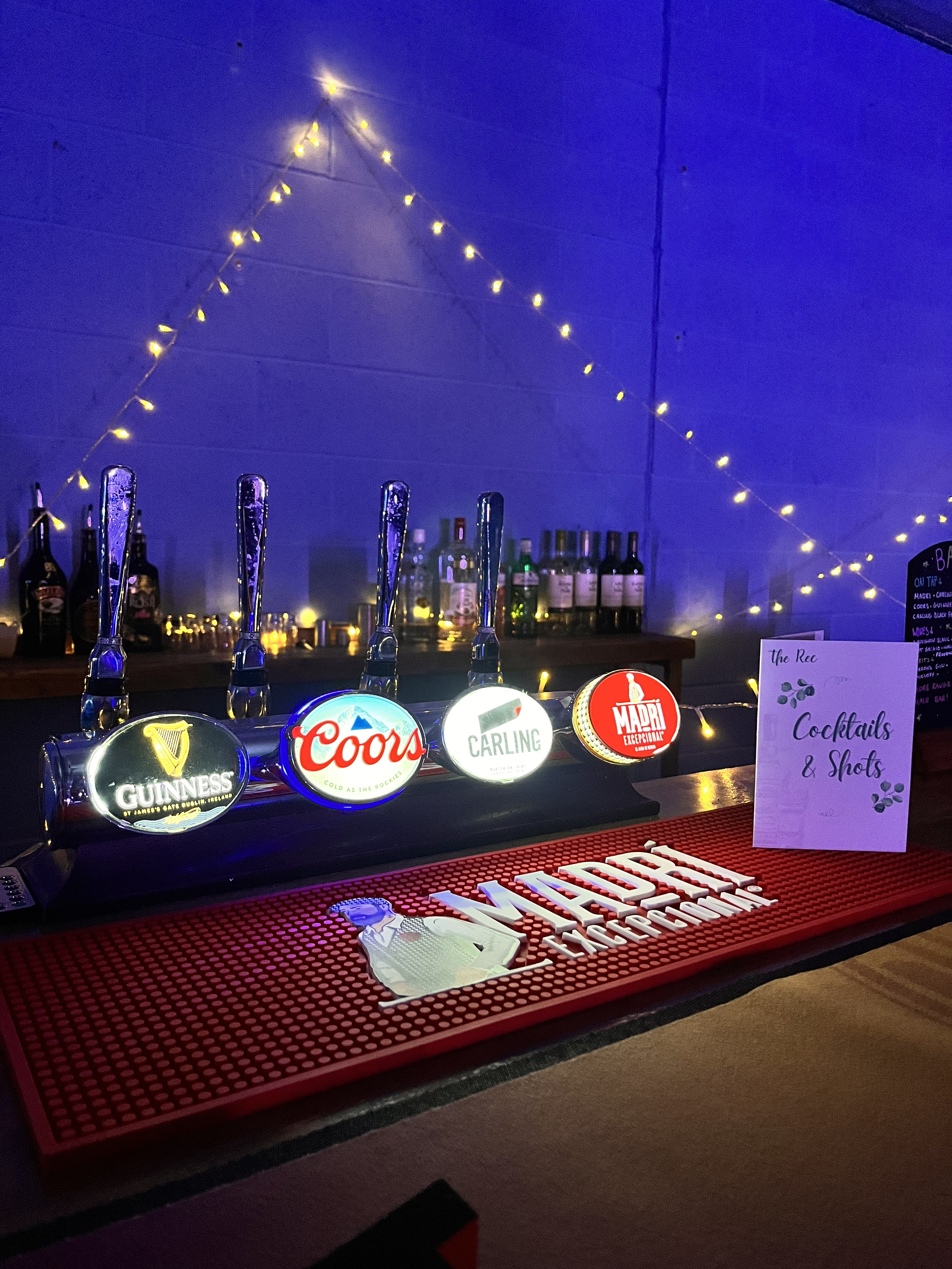 A bar counter with illuminated beer tap handles featuring Guinness, Coors, Carling, and Mâcon logos, lit by string lights. Behind the bar are various liquor bottles. A sign to the right reads 'Cocktails & Shots' with decorative leaf designs.