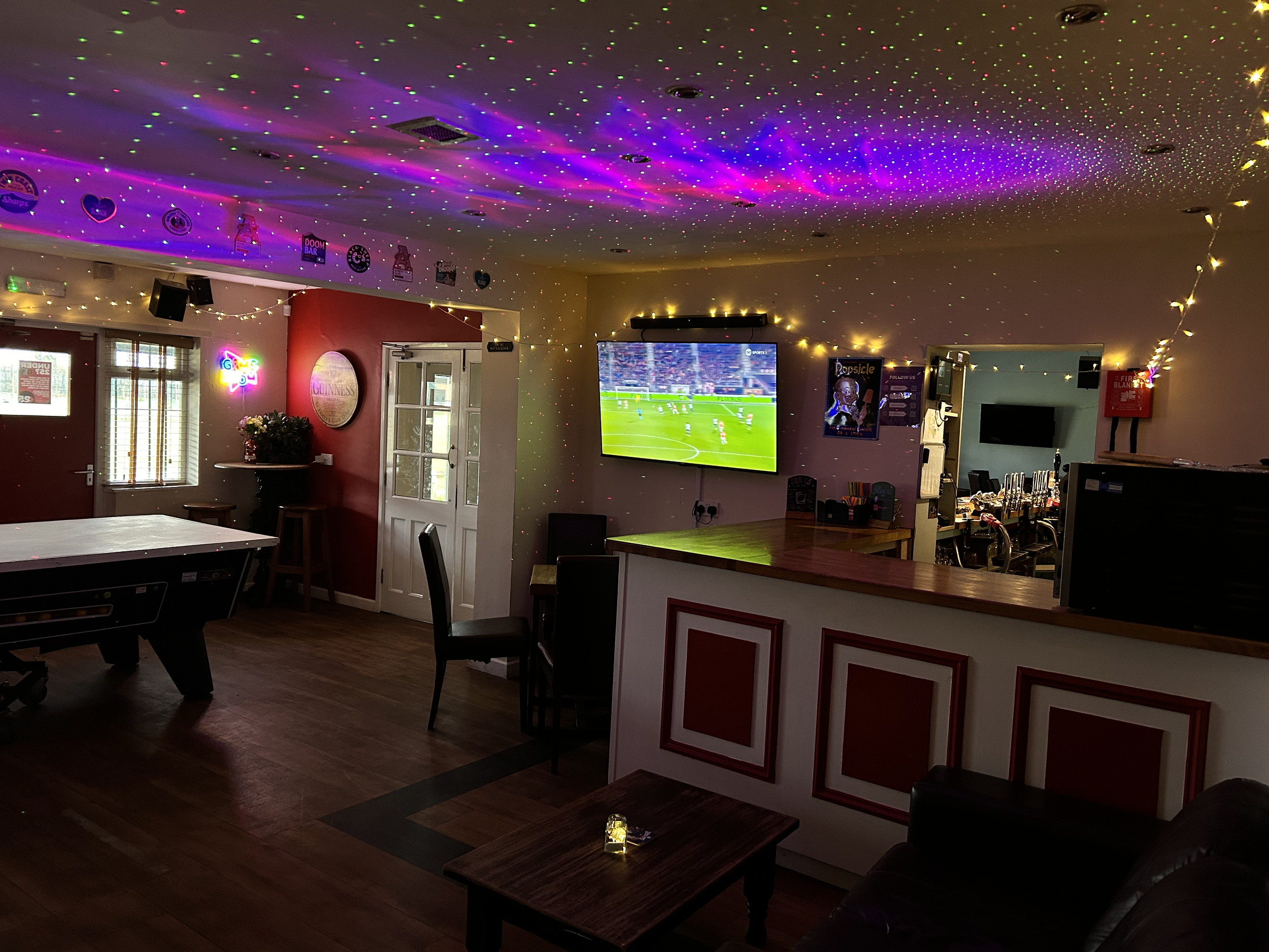Interior of a bar or pub with colorful laser light projections on the ceiling, TV showing a sports game, pool table, and bar with stools.