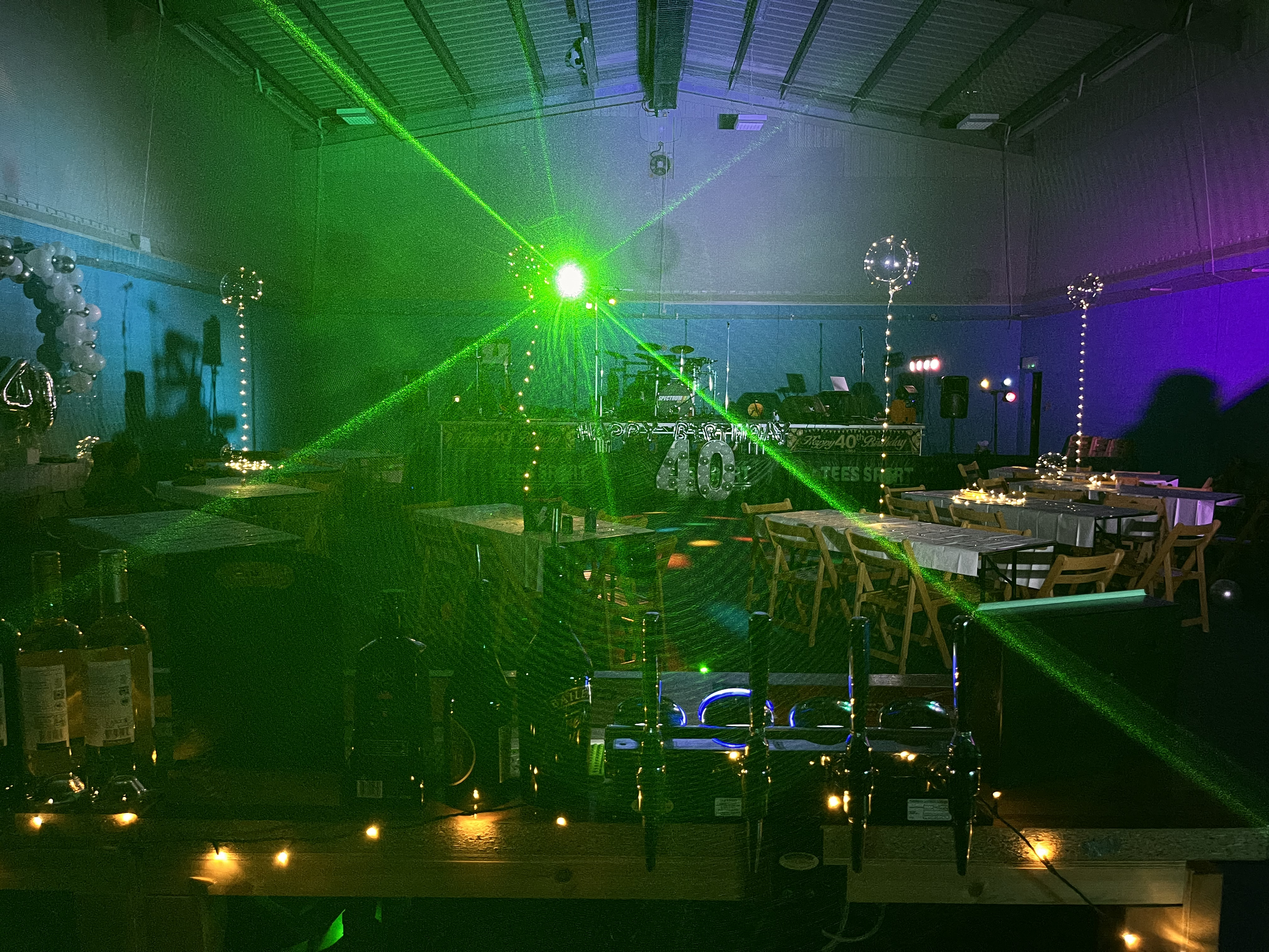 Party setup with tables, chairs, balloons, and a DJ booth illuminated by colorful lighting and green laser lights.