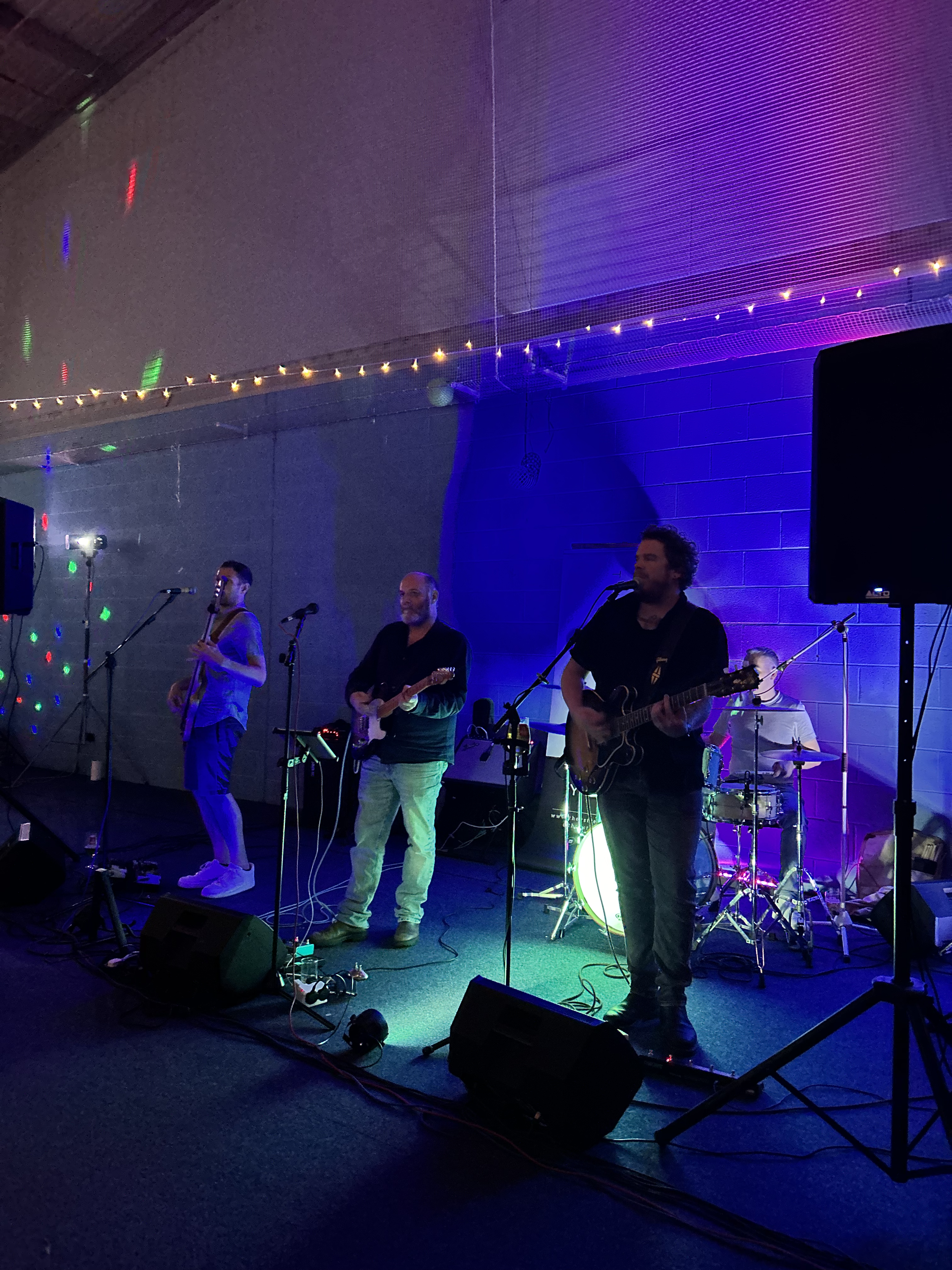 A band of four musicians playing on a stage with colorful lights and a purple background.