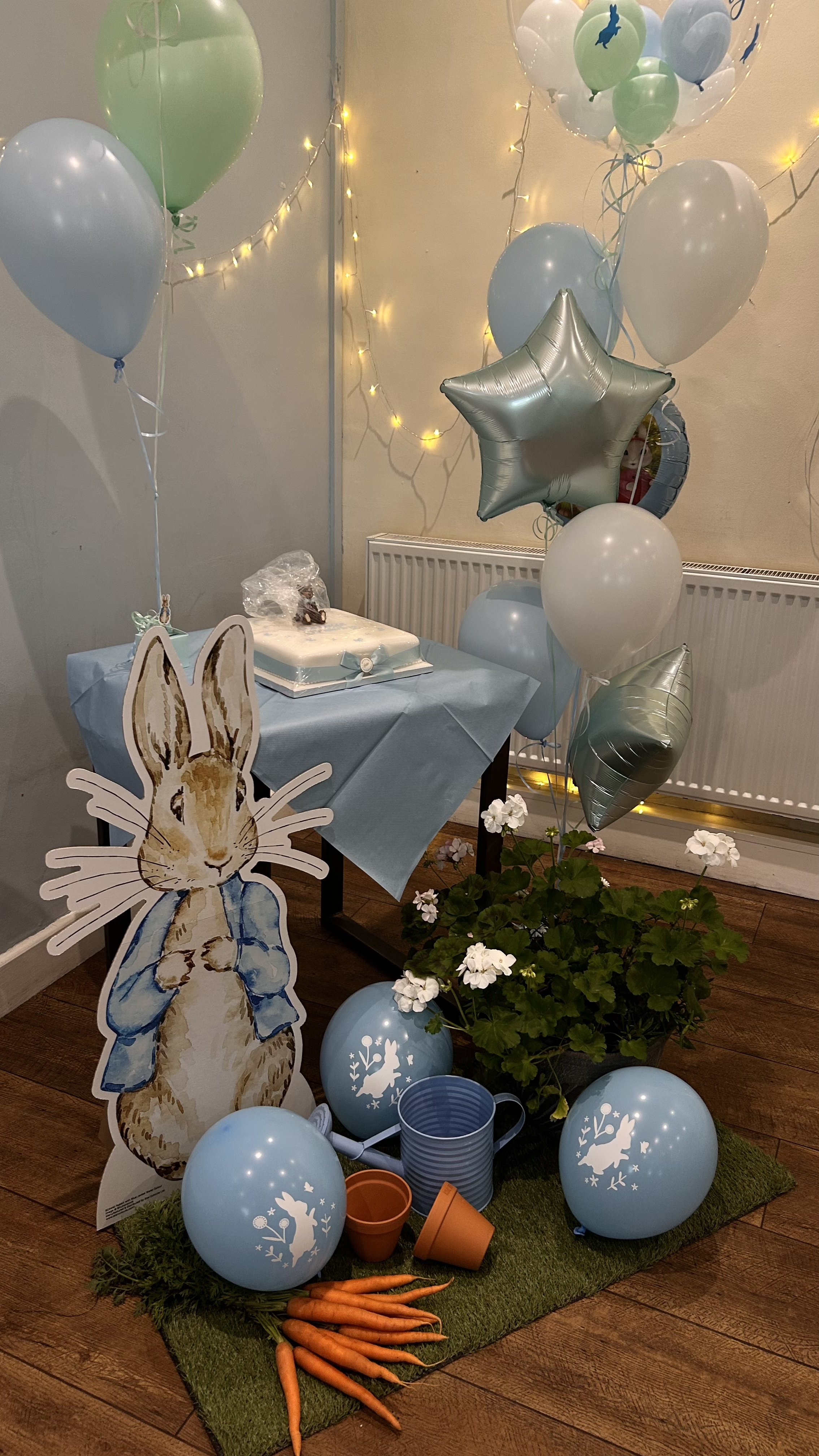 Decorative setup with pastel-colored balloons in green, blue, and silver, some with rabbit designs, along with a cardboard cutout of a rabbit wearing a blue jacket, a potted plant with white flowers, a table with a cake, gardening tools, carrots, and