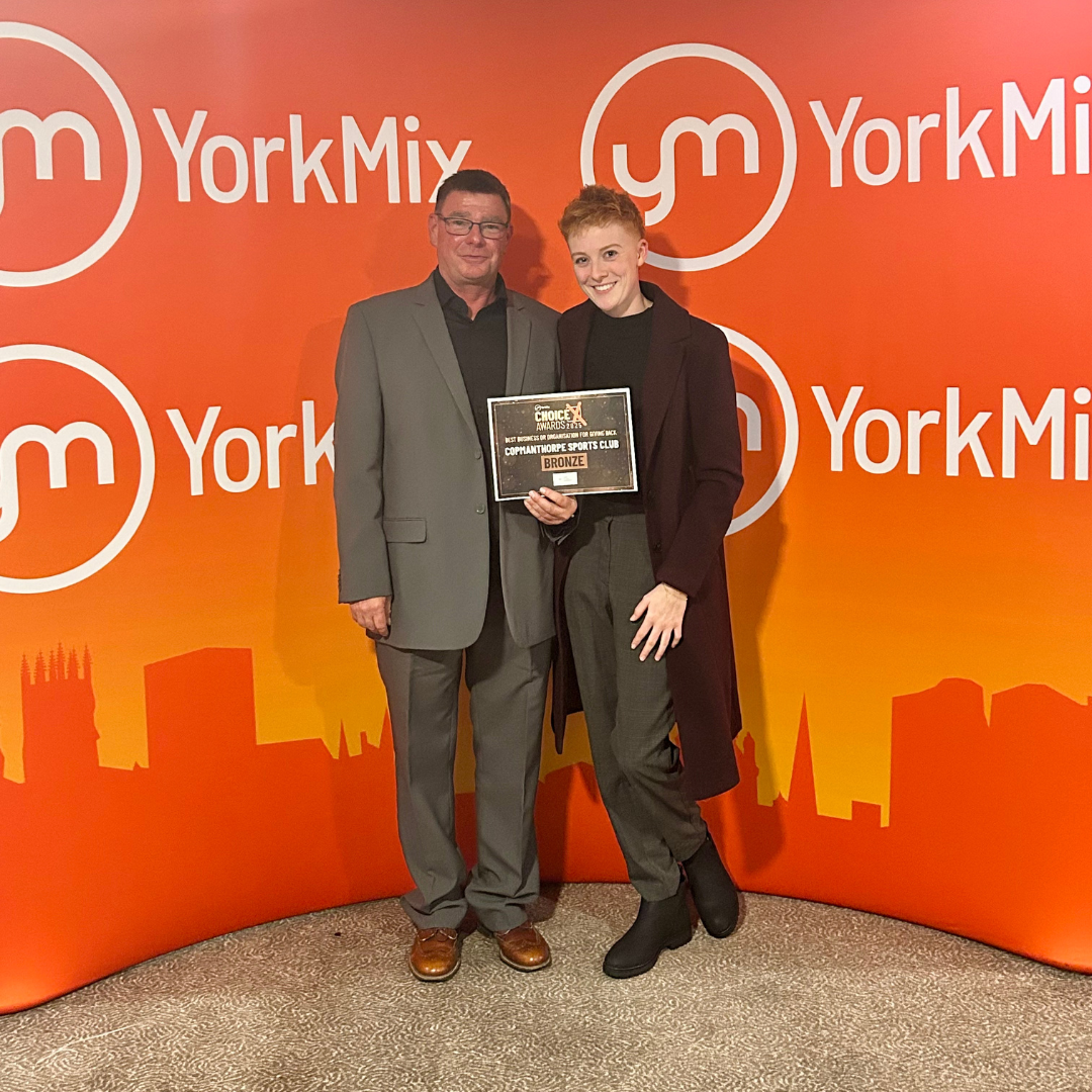 Two people standing in front of an orange backdrop with the YorkMix logo, one of them holding a bronze award plaque for community sports club.