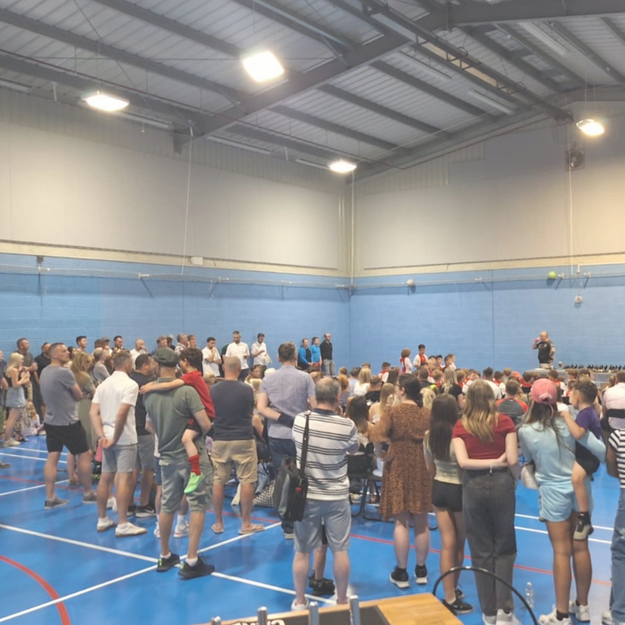 A large group of people inside a gymnasium with blue walls, gathered around a speaker at the front, some standing and others sitting, while listening to a presentation or event.