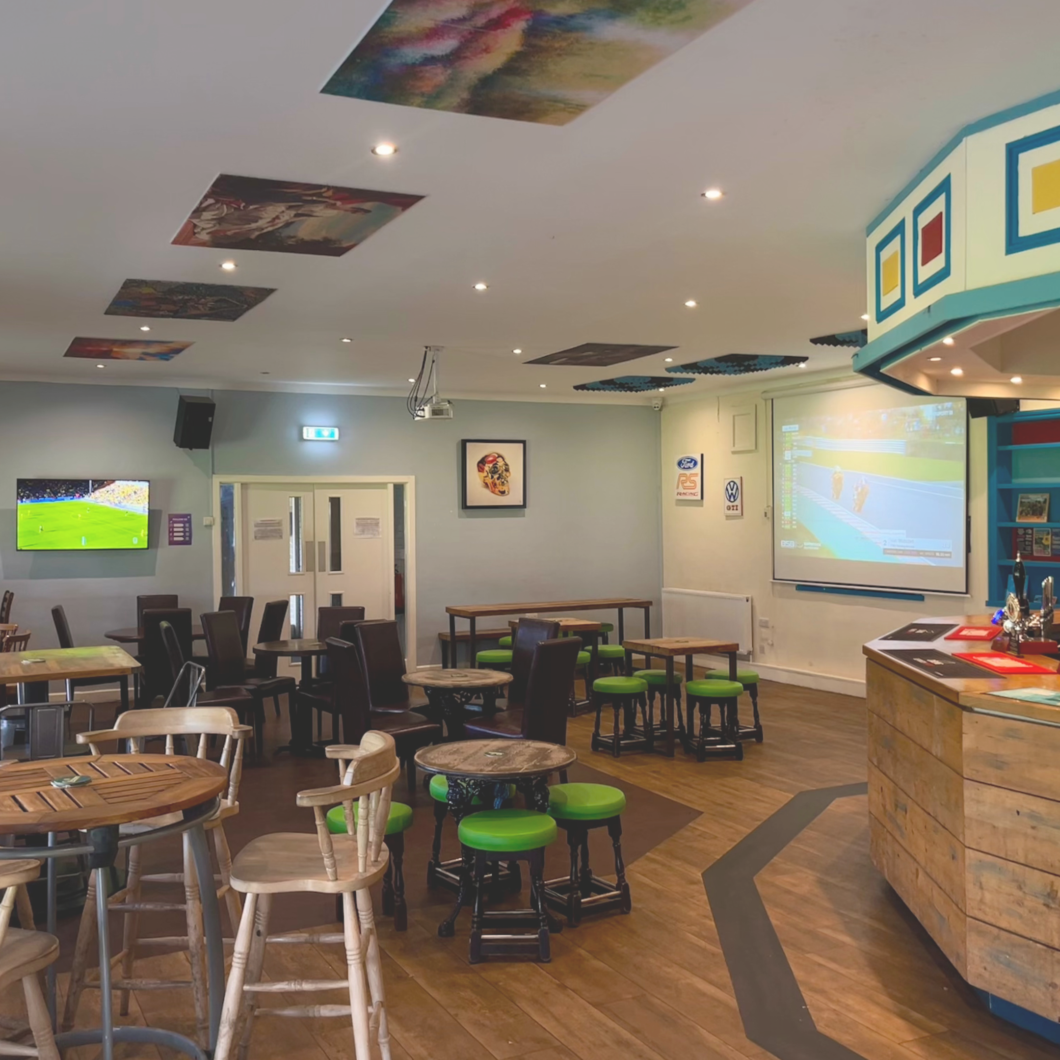 Interior of a sports bar with tables and chairs, a large screen showing a race, a smaller TV showing a soccer game, and sports artwork on the walls.