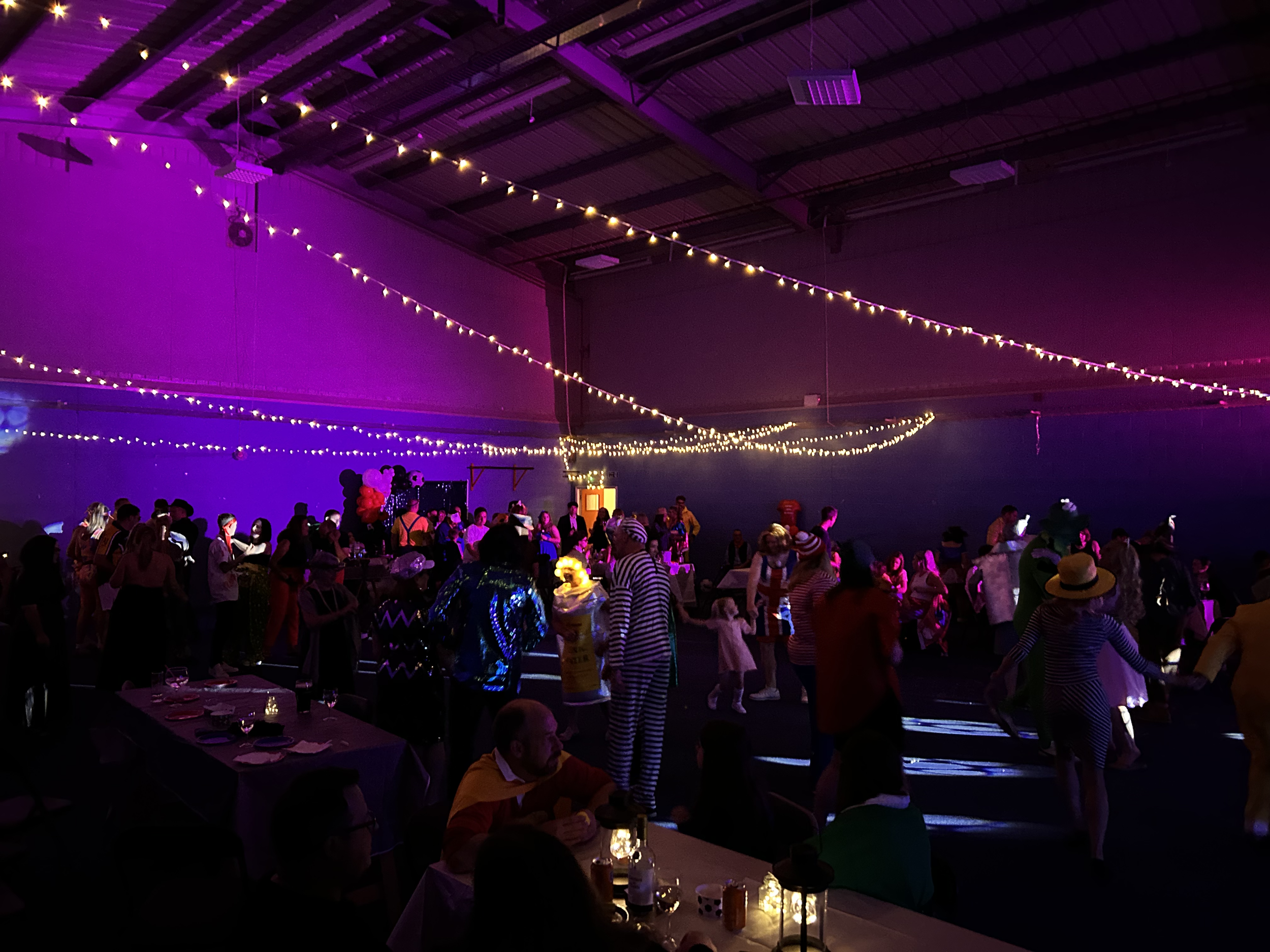 People dressed in colorful costumes dancing and socializing in a dimly lit venue decorated with string lights, balloons, and purple and yellow lighting effects, suggesting a festive event or party.