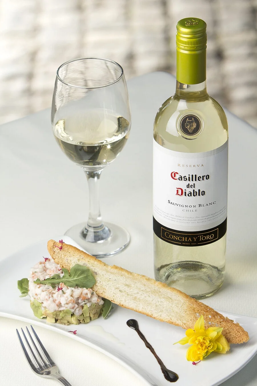 A plate with a seafood salad, a piece of toasted bread, and a yellow flower, along with a glass of white wine and a bottle of Casillero del Diablo Sauvignon Blanc wine.