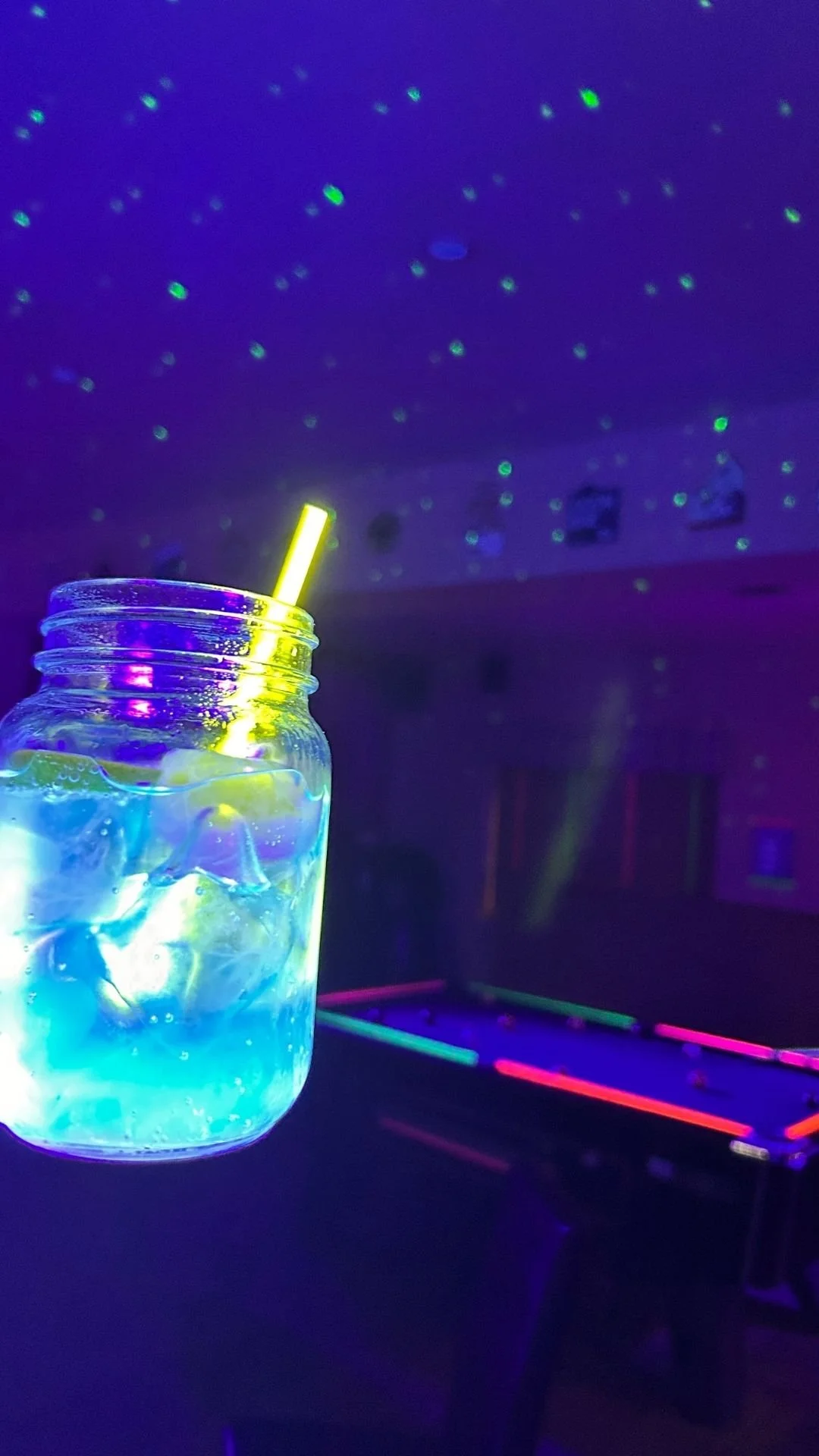 Glass jar with ice and glowing liquid, illuminated under black light with starry effects on the ceiling, and a glow-in-the-dark air hockey table in the background.
