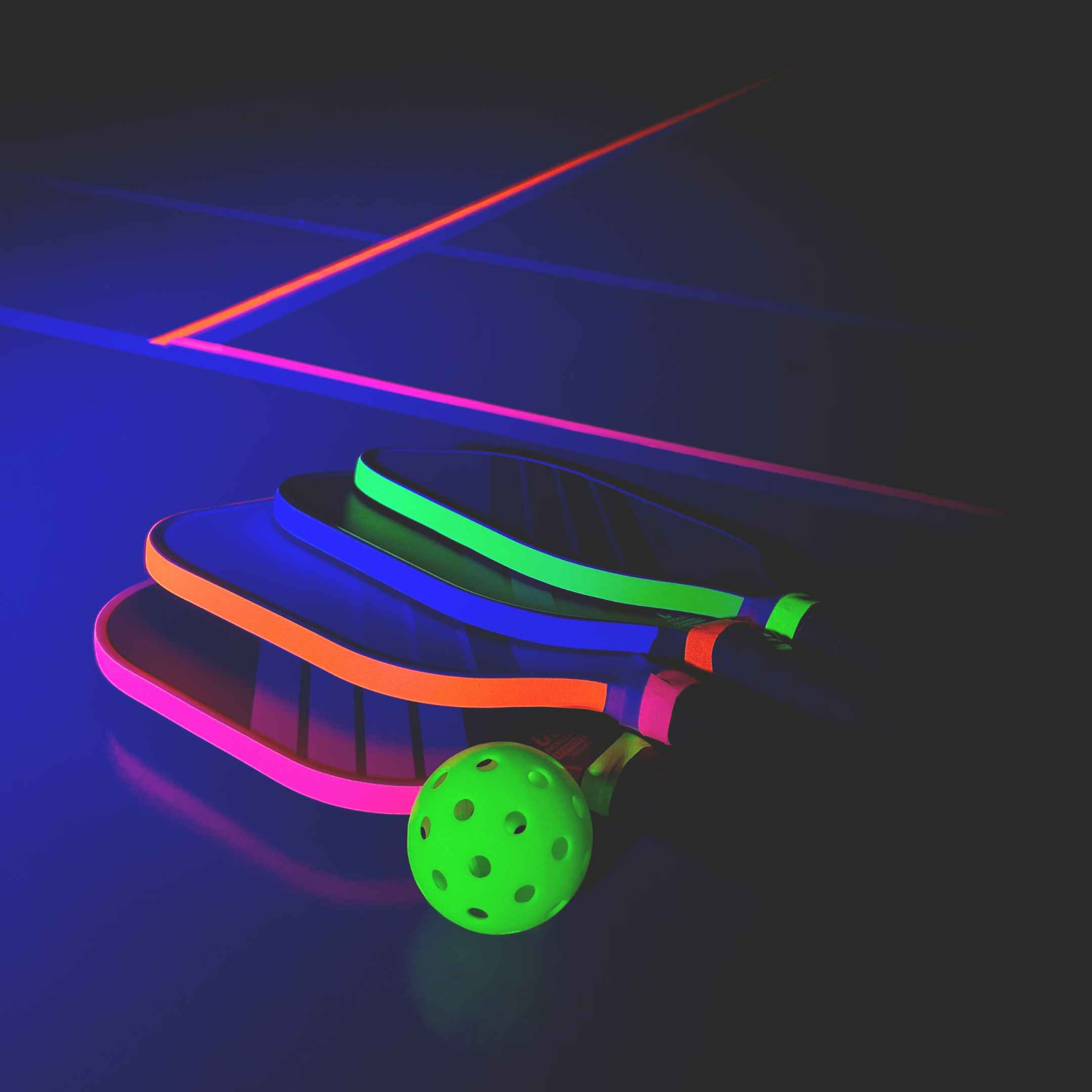 Four neon-colored pickleball paddles with matching neon ball on a dark surface, illuminated by glowing light lines.
